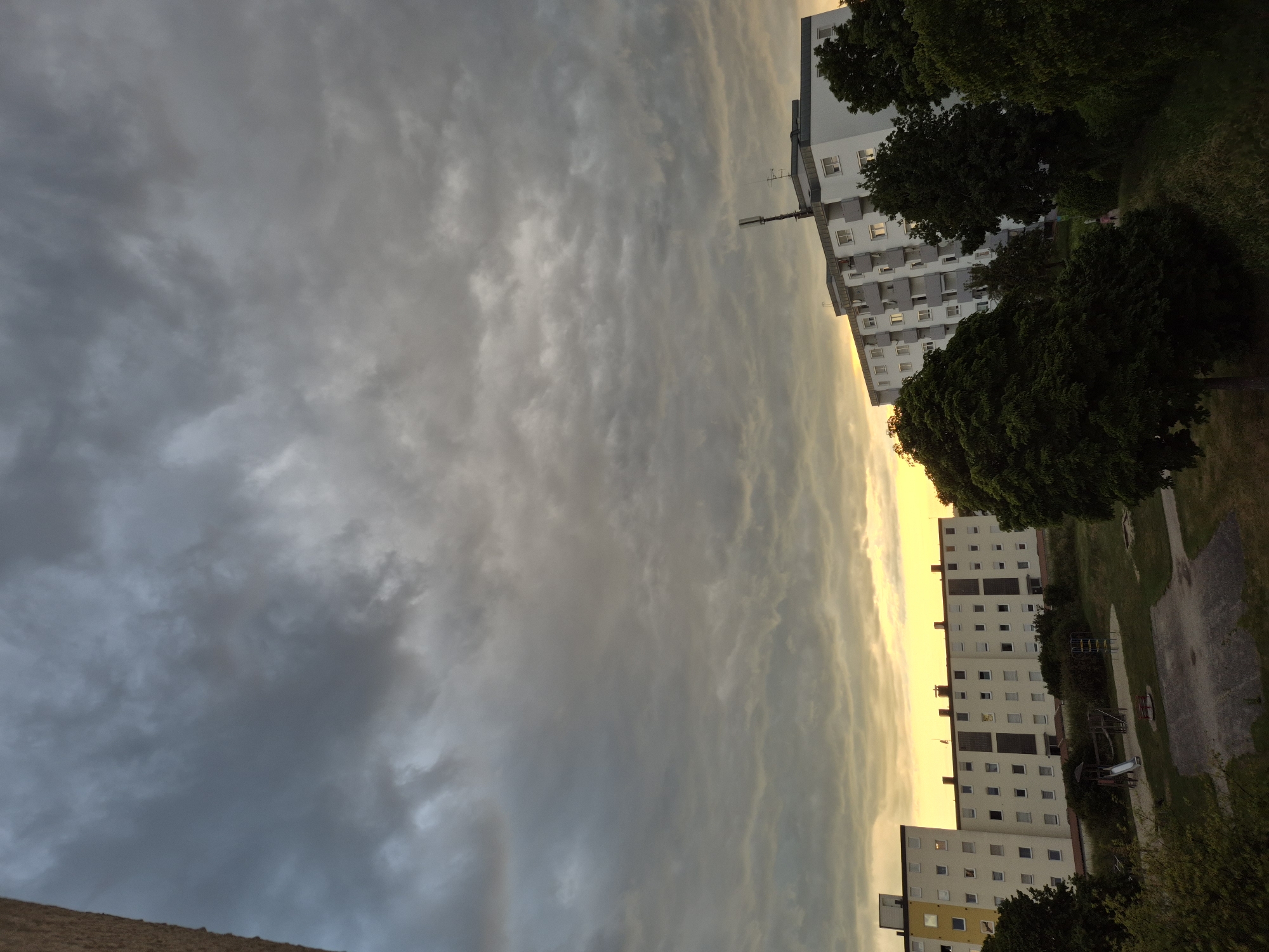 This image captures a residential urban landscape at sunset or sunrise under an overcast sky. The sky is largely covered by a dense layer of grey stratocumulus or altocumulus clouds, with a prominent strip of warm yellow-orange light visible along the horizon, particularly in the lower sky.
The scene features multiple multi-story apartment buildings. On the far left, a portion of a light-colored, multi-windowed building is visible. In the center-left, a long, white, rectangular apartment block with approximately five stories and numerous evenly spaced windows is present. Its flat roof has dark elements, possibly vents or chimneys. To the right, another multi-story, white apartment building features balconies and a prominent, tall communications antenna structure mounted on its roof. The windows of these buildings reflect some of the yellowish light from the horizon.
In the foreground and middle ground, a green communal area or park is visible. This space contains a small playground equipped with a slide, climbing frames, and a sandpit. The ground is covered with grass, and several mature trees with dense green foliage are distributed throughout the area, some partially obscuring the buildings. A paved surface, possibly a path or small court, is also visible near the playground. No discernible people or specific activities are observed. The architectural style suggests a functional residential complex.
The location is an unidentified residential area within the city of Unknown, Unknown.