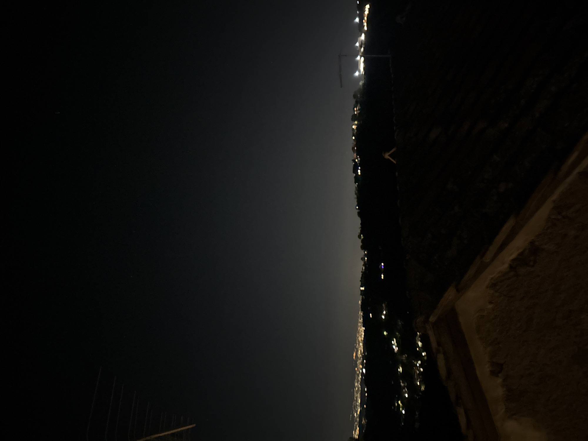 The image shows a night view of Castelnuovo di Porto, Italy, captured from an elevated position, possibly a building or hillside overlooking the town. The foreground is dark and out of focus, with a textured surface (possibly a wall or rooftop) visible in the bottom right. The left side of the frame displays the night sky, with minimal visible stars. The lights of Castelnuovo di Porto stretch horizontally across the middle of the image. A crane structure can be seen among the lights. A dark slope with some foliage outlines the town.