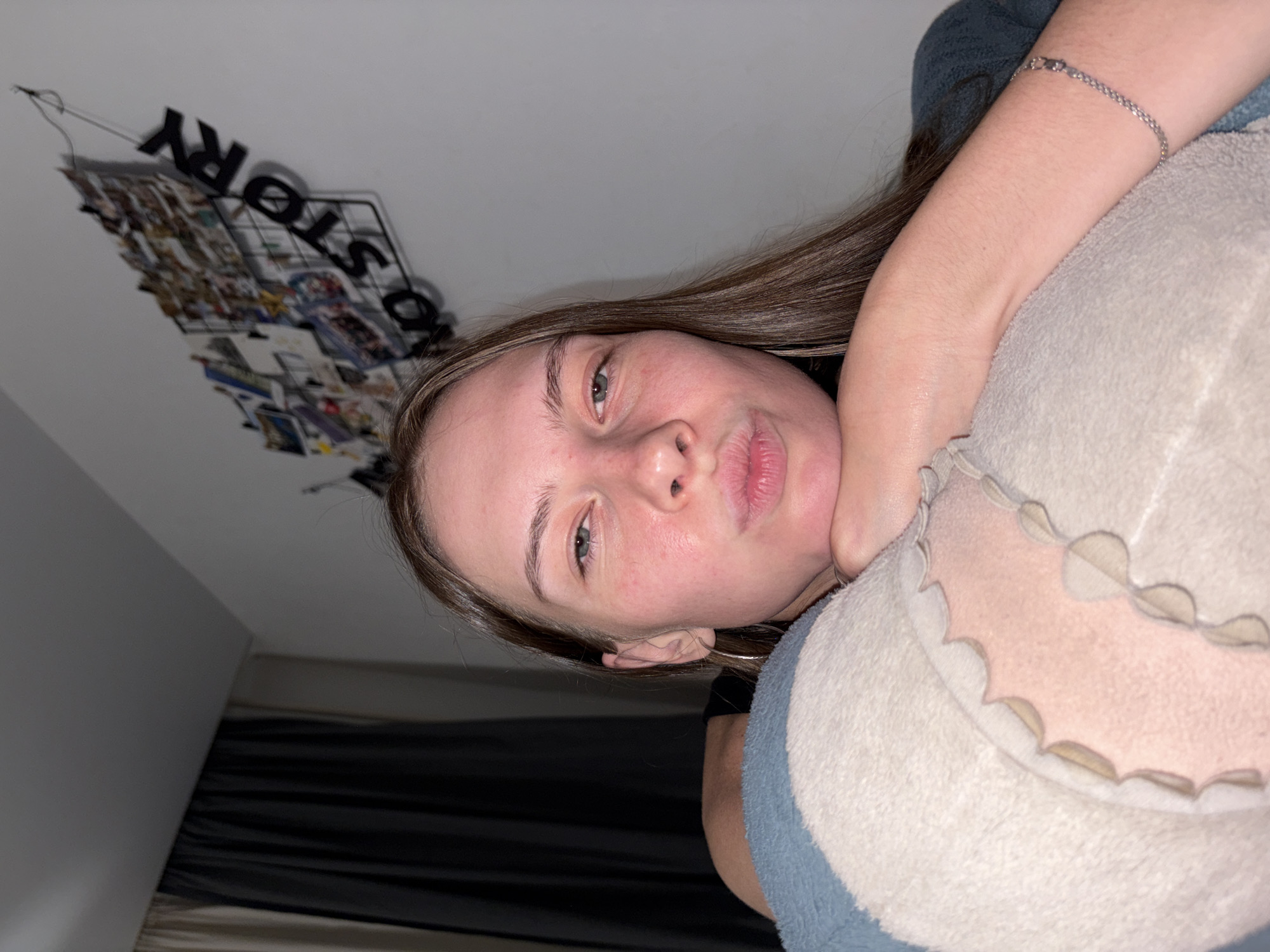 In a dimly lit bedroom, a young Caucasian woman with light brown hair is positioned in the foreground, resting her chin on a plush, light-colored pillow adorned with a scalloped trim. She is wearing a dark blue shirt and a silver bracelet on her right wrist. Her lips are pursed as if blowing a kiss. Behind her, a wire grid is mounted on a light-colored wall, decorated with numerous photographs and the black lettering "NO STORY." To the left of the frame, dark curtains hang, obscuring the view outside. The overall impression is of a personal, intimate space. The provided image does not contain any identifiable information that would allow for specific location details within Perm, Russia.