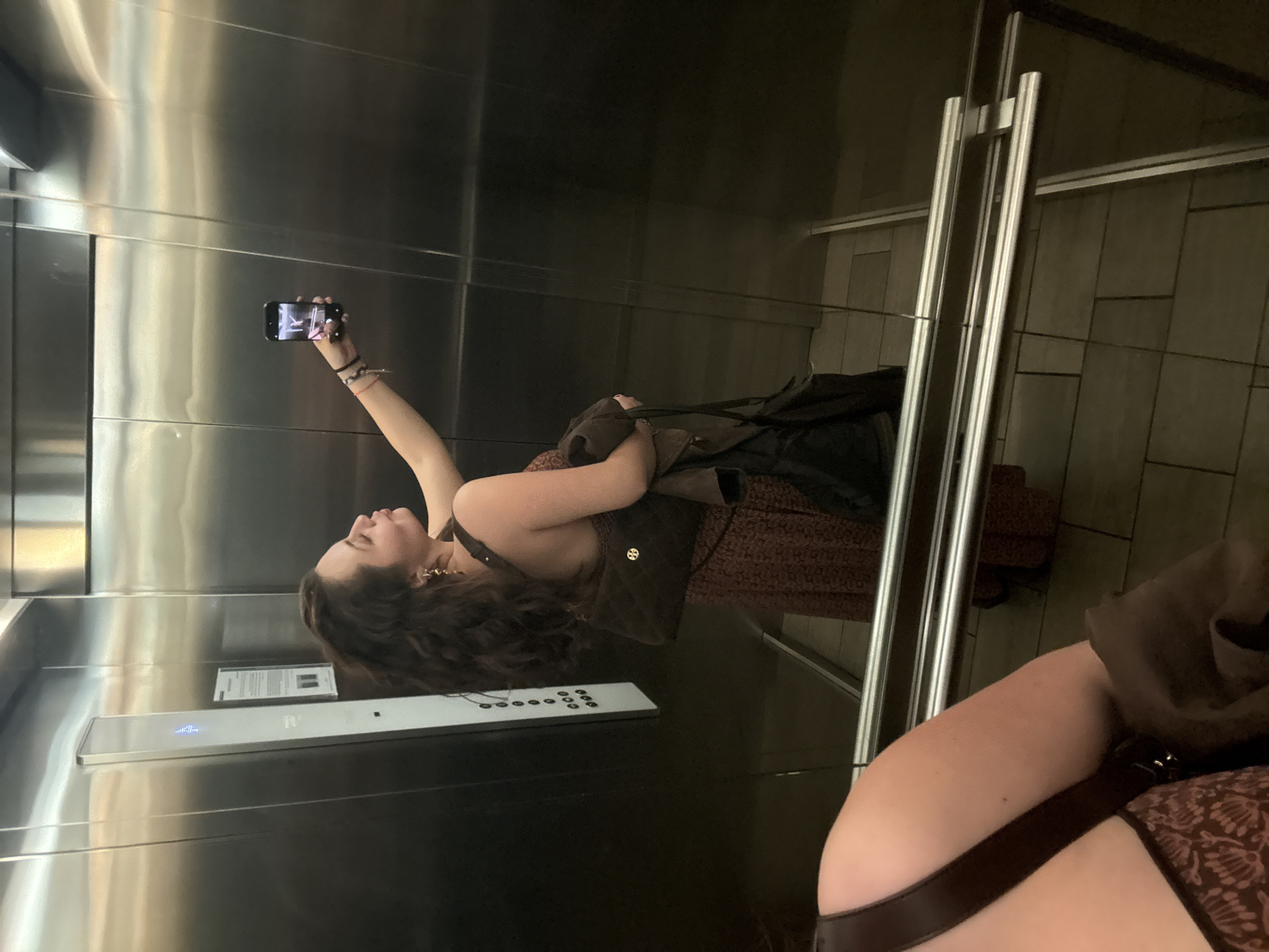 A young woman is taking a selfie in an elevator, likely in Mexico City, Mexico. She is holding a smartphone up with her right hand, her arm extended towards the top left corner of the frame. The elevator has shiny metal walls and a control panel with buttons. A closed glass door is visible on the right side of the frame. Another person is partially visible in the lower right corner, with their arm and shoulder in view, wearing a brown patterned dress. The lighting is bright and artificial, reflecting off the metal surfaces of the elevator. The woman taking the selfie is wearing a brown dress and a brown bag.