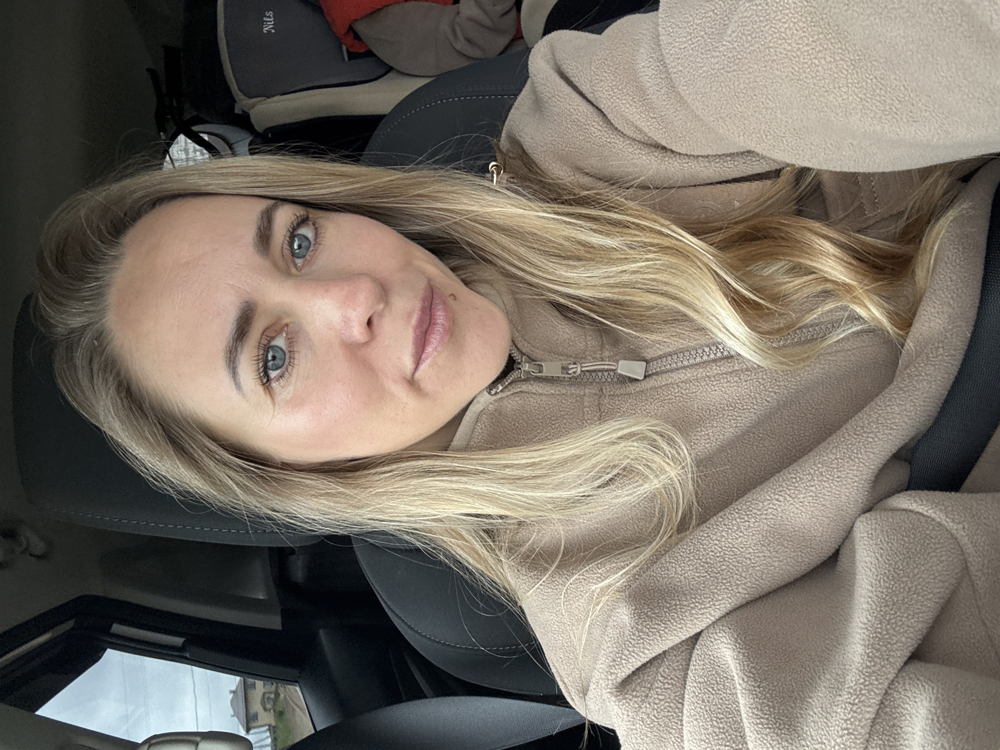 A woman with long, blonde hair and blue eyes is seated in a vehicle. She is wearing a beige fleece jacket with a zipper. A seatbelt is visible across her body. The interior of the car is gray and black. Outside the car window, a glimpse of a building and overcast sky can be seen. The location cannot be determined to be in Chaikovsky, Russia with the information provided.