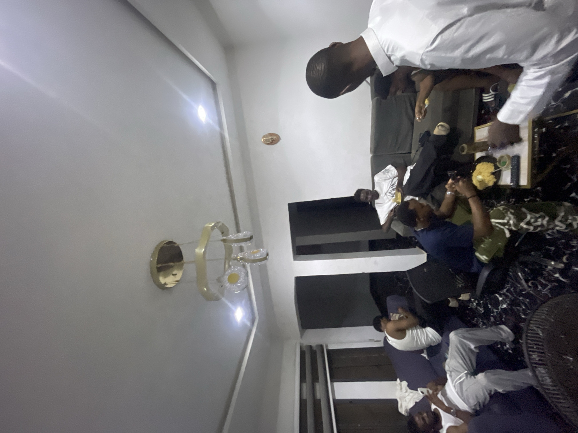 The image appears to be taken indoors, possibly in a living room in Abuja, Nigeria. Several people are present. Some are sitting on a sofa while others are seated in chairs or lying down on the floor. There are items on the table such as food and remote controls. A lighting fixture is seen on the wall. The setting suggests a casual gathering or social event.