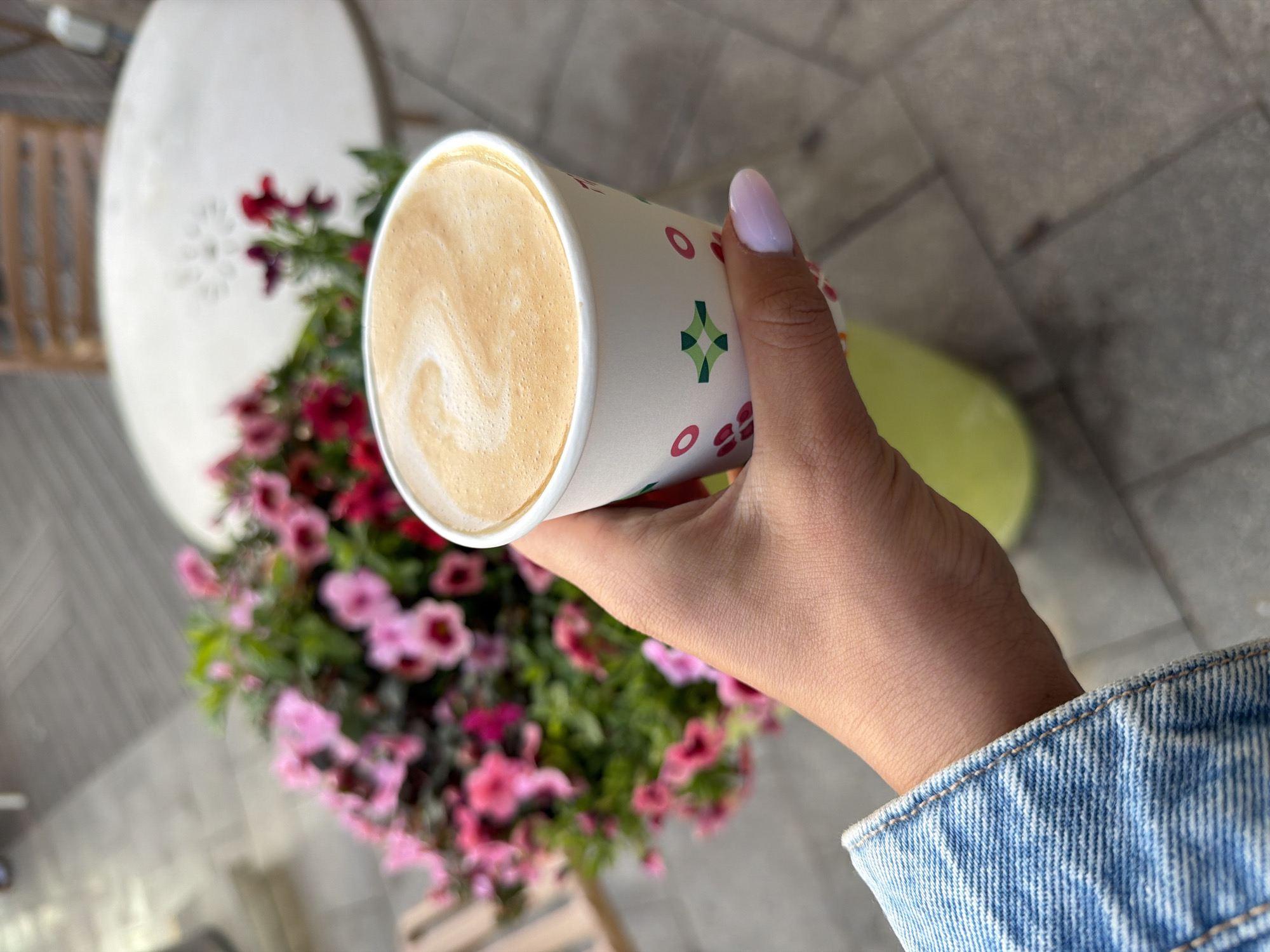 A medium shot depicts a person holding a cup of coffee in front of a flower arrangement.
The cup, held in a right hand with a light purple fingernail, is white with a pattern of green and red shapes. The coffee inside is light brown with a swirling design on top. The person is wearing a light-wash denim jacket.
Behind the cup and hand is a blurred background consisting of a round white table with a floral design, a collection of pink and red flowers, and a light green object on a gray-tiled surface. The scene is presumed to be in Tel Aviv, Israel.