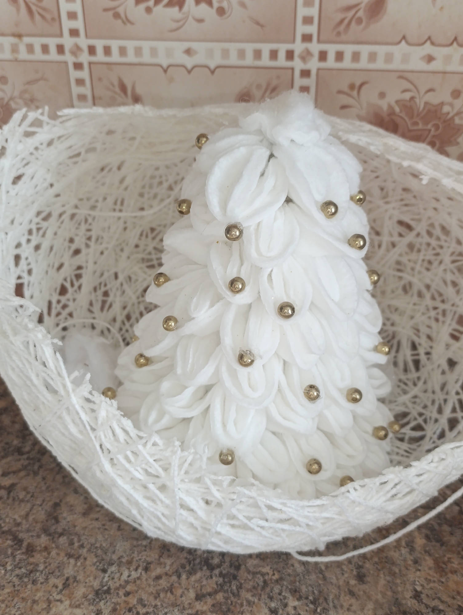 This is a close-up, eye-level, indoor shot of a decorative white Christmas tree. The tree is small and conical, adorned with small, gold beads. It is nestled inside a larger, open, white woven basket. The background is a textured, tiled wall with a subtle, floral pattern. The surface beneath the basket is a mottled, brown and grey speckled material, possibly a countertop. The lighting appears to be soft and diffused, suggesting it might be daytime with indirect light or artificial indoor lighting. There is no visible text in the image. The overall impression is of a festive, handmade decoration.