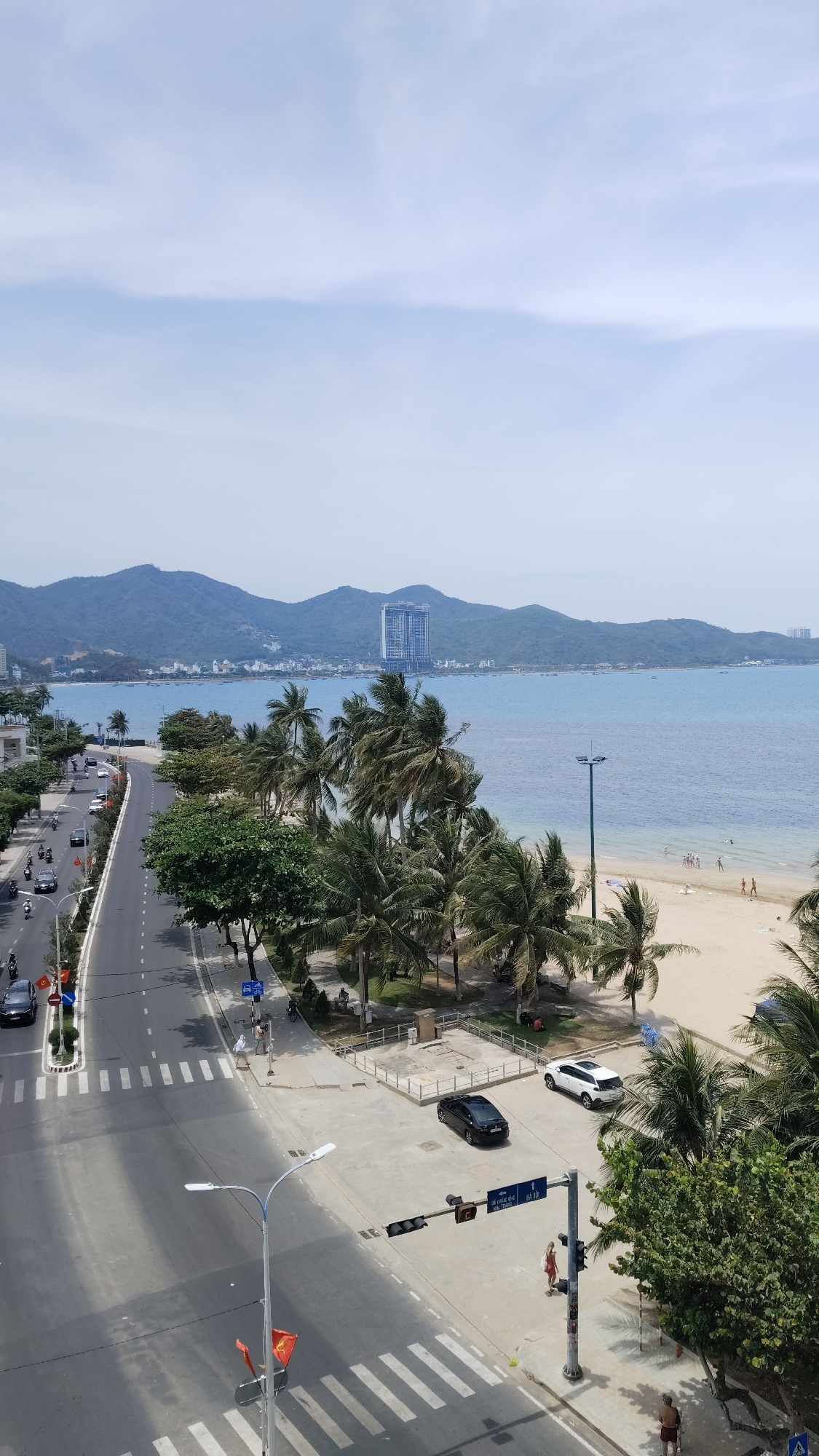 This is a wide-angle, daytime shot of a coastal scene, presumably in Bac Nha Trang Ward, Vietnam. The view is elevated, looking down on a multi-lane road running along the coastline. On the left side of the road, there is traffic including cars and scooters moving along the street. Sidewalks line the road with occasional pedestrian traffic. A row of trees and vegetation separates the road from the beach area. The beach stretches to the right side of the frame, with a sandy expanse and several people present near the shoreline. The water appears calm with boats visible in the distance. A tall building is visible on the coastline. The sky is overcast with bright areas. Traffic lights are visible at a crosswalk with pedestrians crossing.