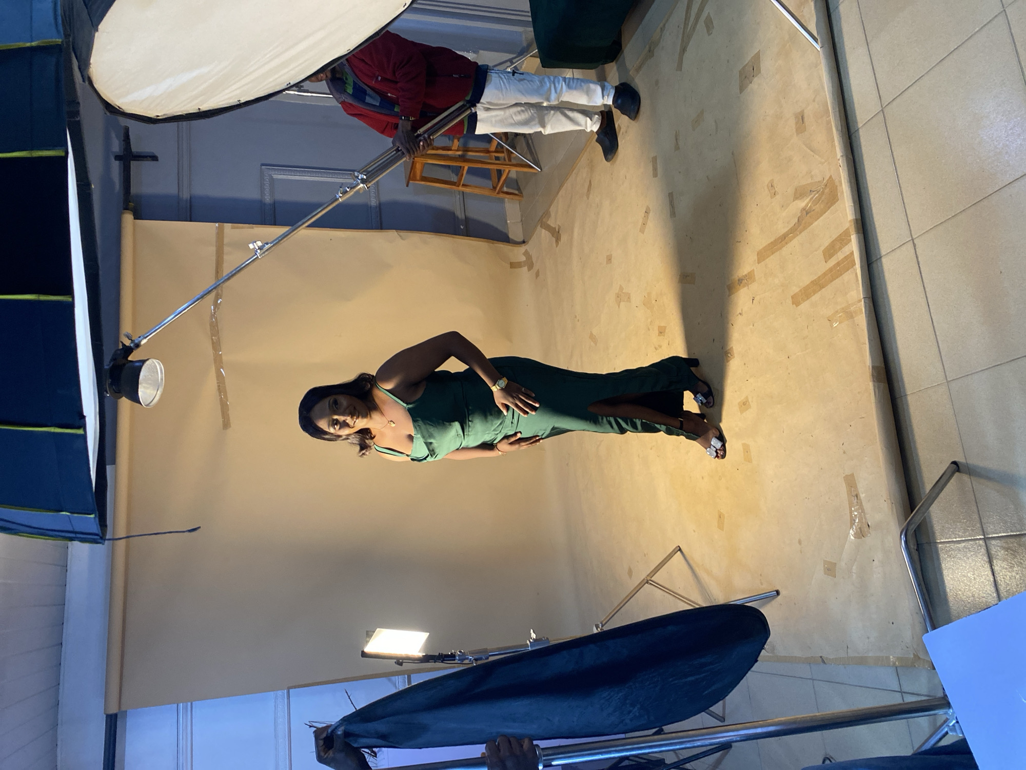 In a studio setting, a Black woman stands center, wearing a floor-length, emerald green cowl neck dress with a thigh-high slit, and silver strappy heels. She has dark, shoulder-length hair and is adorned with a gold necklace and wristwatch. The woman is posing with one hand on her hip.
The studio has a light brown backdrop, a tiled floor, and is equipped with professional photography lighting and equipment. To the left, a black fabric is held by a person, partially obscuring a bright light stand. To the right, a person in a red jacket, blue and green striped shirt, white pants, and black shoes stands near a large softbox. Another person is visible in the far background, and a wooden stool is positioned behind the woman on the right.
The image was taken in the city of Bukuru, Nigeria, within a photography studio. The scene depicts a professional photoshoot in progress.