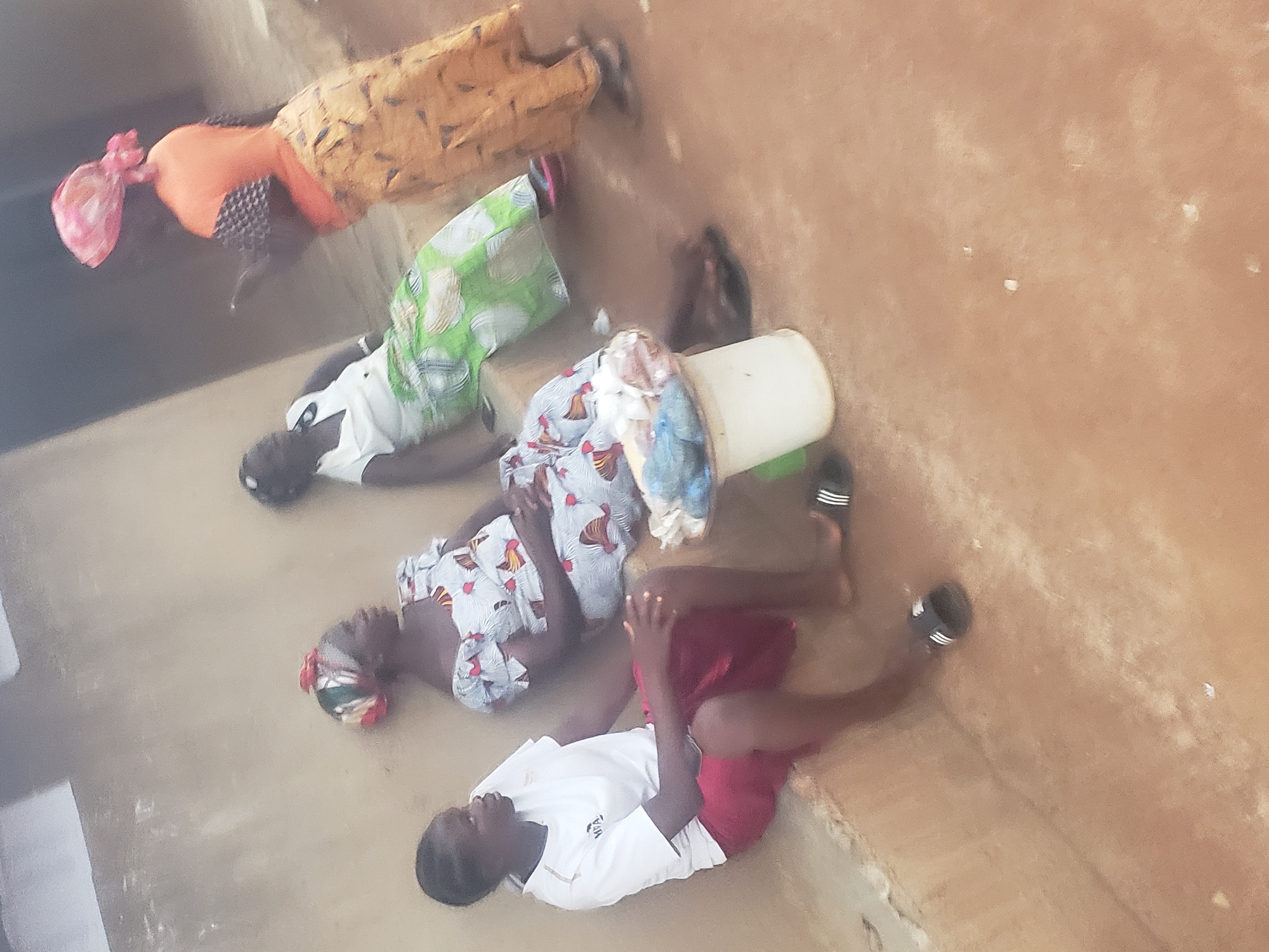 In Kachia, Nigeria, four women are gathered outdoors, near a weathered, light-colored wall. Two women are seated on a low, concrete ledge, facing left. The woman on the left wears a white t-shirt with a logo and a red skirt. The woman next to her is dressed in a patterned blue and white dress and is looking downwards. To their right, another woman is standing and facing them, wearing a white top and a green and white patterned skirt. She appears to be in conversation. Further to the right and slightly behind her, a fourth woman is visible from the back, wearing a bright orange top and a patterned yellow skirt. She has a pink headscarf and is looking to her right, possibly engaged in the same conversation. In front of the seated women, a small white bucket sits on the ground, filled with what appears to be various items wrapped in plastic. The ground is a dusty, reddish-brown earth. Several pairs of sandals are scattered around the area.