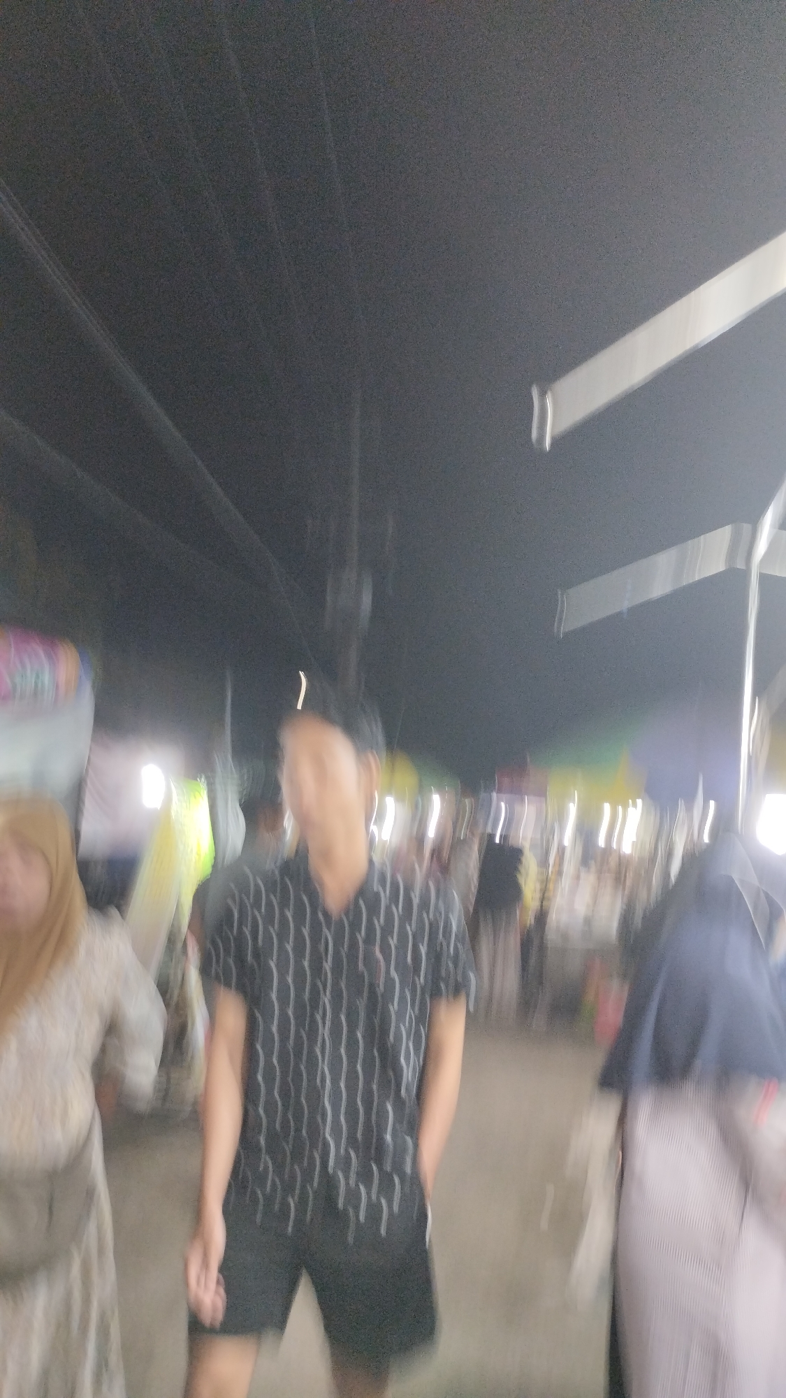 The image depicts a street scene at night, likely in Sambungrejo, Indonesia, characterized by the presence of people and various stalls or vendors, suggesting a market or festival atmosphere. The lighting is dim and appears to be artificial, with some sources casting bright, elongated streaks due to motion blur.
The central subject is a person wearing a short-sleeved shirt with a vertical striped pattern in black and white, and black shorts. To the left of this individual, another person is partially visible, wearing a light-colored outfit and a headscarf. To the right, another person is seen wearing a dark hijab and a striped skirt. The overall image suffers from significant motion blur, making it difficult to discern fine details or facial expressions.
In the background, there are indications of stalls or structures with lights, and the presence of other blurred figures suggest a gathering of people. Overhead, what appear to be electrical wires are visible against the dark sky. Two white, elongated shapes, possibly part of a structure or signage, are also prominent in the upper portion of the image. The time of day is clearly night, and there are no immediate cues about the weather, though the low lighting might suggest it is not raining. The scene conveys a sense of activity and public gathering, typical of a night market or local event. No visible text is discernible due to the blurriness.