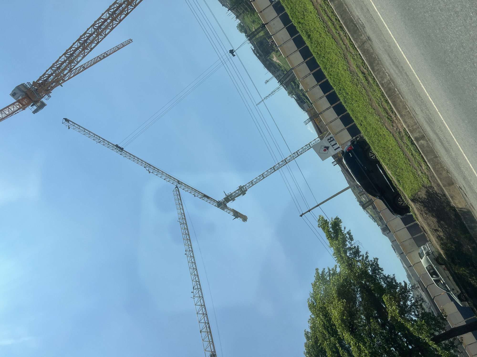 This image captures a construction scene in Jiru, Nigeria, under a clear, bright blue sky. Four yellow lattice-boom tower cranes are prominently featured against the sky, indicating active construction. One crane is in the upper right, displaying its machinery housing and operator's cab; another extends horizontally across the mid-left; a third extends downwards in the middle-right; and a fourth is partially visible on the far left. Below the cranes, a segmented construction fence with light-colored panels and a dark base separates a construction site from a paved road. Behind the fence, a white sign with a red circular logo and the black text "ITB" is partially visible. Further back, a rising landmass or hill shows visible concrete structures, suggesting ongoing building development. A strip of vibrant green grass runs along the roadside, bordered by a concrete curb. A paved road with a white line marking occupies the lower right quadrant. A dark-colored SUV is parked on the grassy verge, partially obscuring the ITB sign, and a lighter-colored sedan is visible further down the road to the left, partially hidden by a large, dense green-leafed tree. Utility poles and power lines traverse the upper and mid-ground. No individuals are discernible in the scene. The image is captured with a noticeable upward and counter-clockwise tilt.