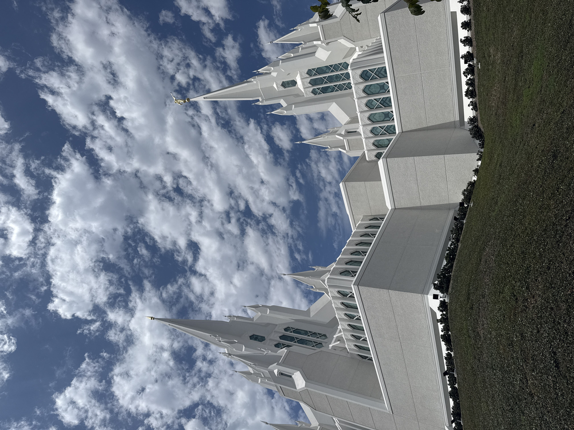 The media file shows a close-up, low-angle view of the San Diego California Temple, a white, multi-spired building against a bright blue sky filled with scattered white clouds. The architecture features numerous sharp spires and arched windows with teal-tinted glass. The building's textured, light gray facade is visible. To the right of the temple, a meticulously maintained green lawn stretches out, bordered by a line of dark green shrubs. The image is taken outdoors during the day, under clear and partly cloudy weather conditions. The perspective emphasizes the grandeur and scale of the temple. The scene is serene and evokes a sense of peace. No people are visible in the frame. There is no visible text.