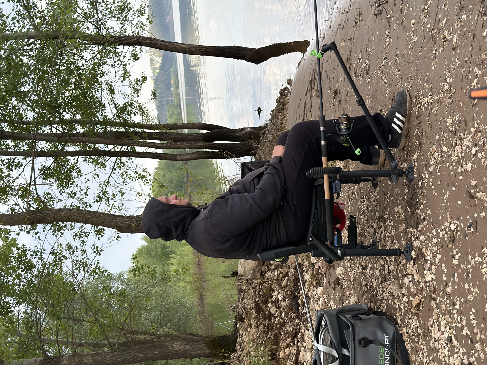 A man sits in a black fishing chair on a muddy bank along the Kama River in the city of Chaikovsky, Russia. He wears a black hoodie and black Adidas sneakers with white stripes. A fishing rod is set up on a stand in front of him, near the water’s edge. A gray and black bag sits beside the chair. Trees line the bank, and the river extends into the distance, showing some buildings across the water. The ground is a mix of mud and small white rocks or shells.