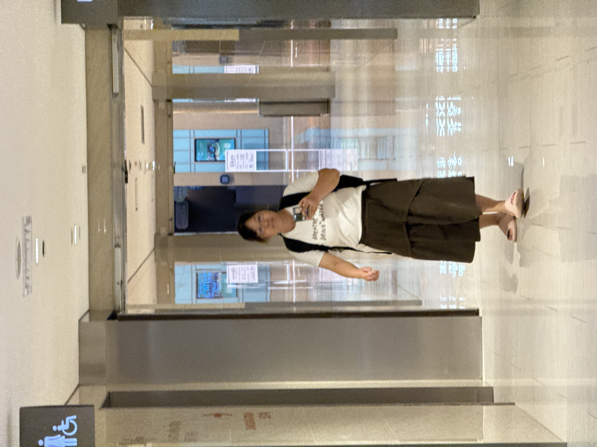 A woman walks down a well-lit, modern hallway with reflective tiled floors. She is wearing a white t-shirt with text on it, a dark brown skirt, and pink sandals. She is holding a smartphone in her right hand and has a black backpack on. To her left, a sign with the international symbols for women and individuals with disabilities is visible on a gray pillar. The hallway appears to be in a public indoor space, possibly a mall or a transportation hub, given the signage and architecture. The reflection on the floor suggests overhead lighting and possibly architectural elements in the distance. The scene is captured from a slightly low angle.