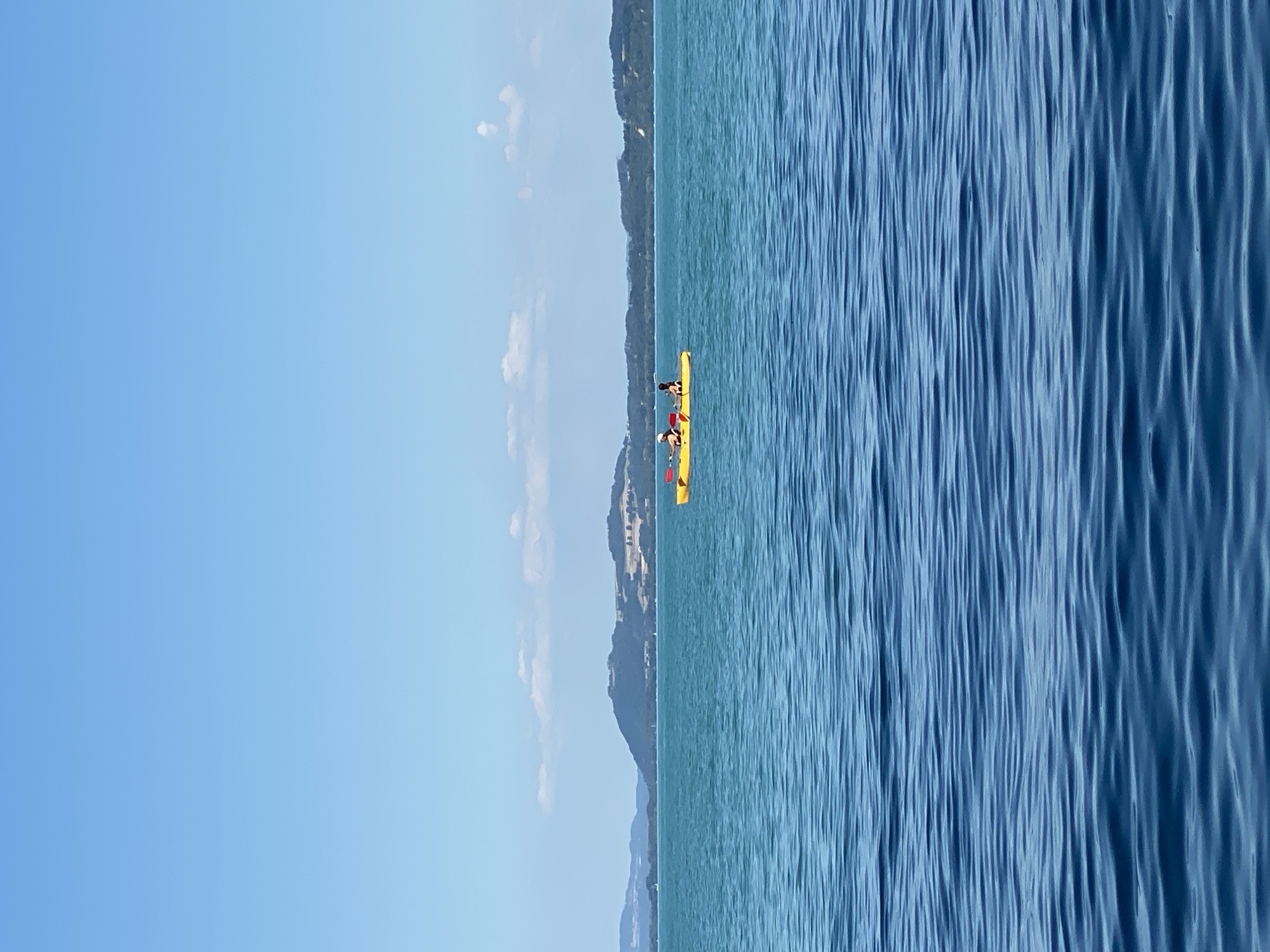 In the city of Cortaillod, Switzerland, on a clear day with a bright blue sky, a yellow kayak carrying two individuals is visible on the turquoise waters of Lake Neuchâtel. The two people are paddling the kayak. In the background, rolling green hills with scattered trees and structures form a distant shoreline. The water has gentle ripples.