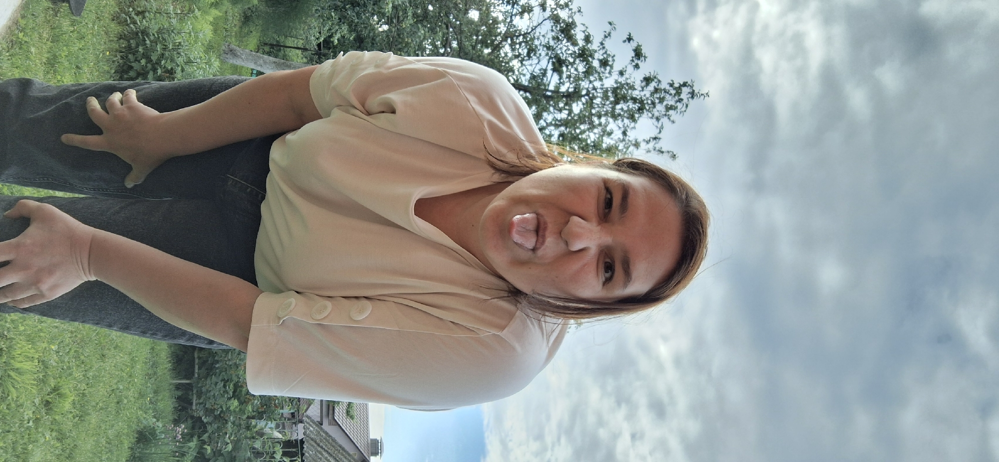 This is a slightly rotated, close-up, outdoor selfie of a woman with light brown hair and brown eyes. She is wearing a light beige t-shirt with decorative buttons on the sleeves and dark denim jeans. The woman is sticking her tongue out at the camera, with a playful or mischievous expression. Her face is the primary focus of the image.
The background includes lush green foliage from trees and grass, suggesting a natural setting. In the lower portion of the image, a glimpse of a roof with tiles can be seen, possibly indicating a residential area or a building within a park. The sky above is cloudy, with a mix of white and grey clouds, hinting at an overcast or partly cloudy day. The lighting suggests it is daytime, likely late afternoon or early evening given the softness of the light. The overall impression is casual and spontaneous, capturing a moment of lightheartedness in an outdoor environment. No text is visible in the image.
