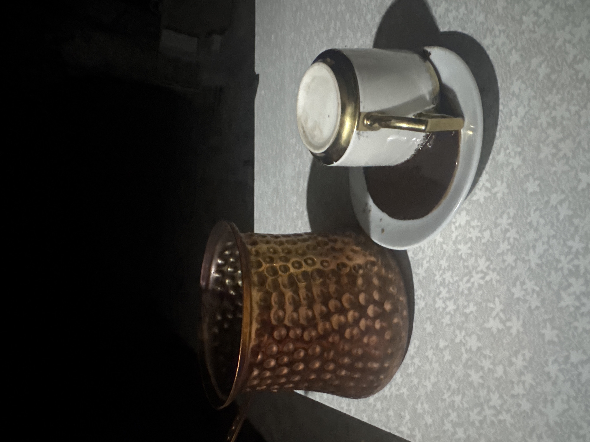 A copper Turkish coffee pot sits to the left of a white coffee cup with a gold rim and handle, which is overturned on a saucer. The cup is filled with dark liquid that has spilled onto the saucer. The objects are placed on a light-colored table with a subtle white floral pattern. The background is dark, suggesting it is nighttime. The scene is located in Taganrog, Russia.