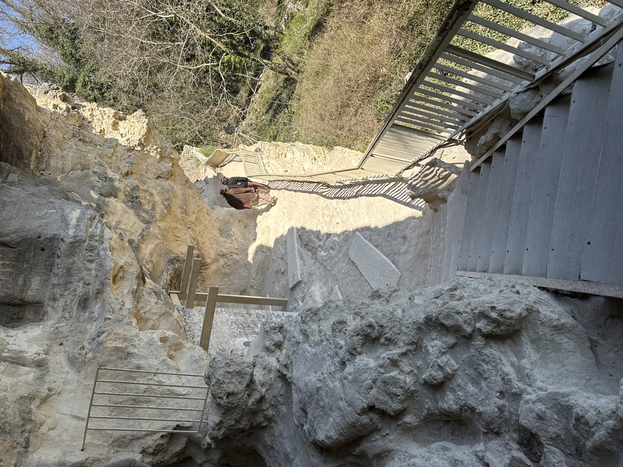 The image depicts a scene within the Aladzha Monastery complex in Varna, Bulgaria. The view is angled downward, showcasing a series of stone steps and walkways carved into a cliff face. A metal staircase runs along the edge of the cliff, providing access to different levels. Below, a platform with wooden beams and a section of cobblestone paving is visible. Further down, a metal railing is mounted to the rock. The surrounding environment consists of exposed rock faces, sparse vegetation, and bare tree branches. Sunlight casts shadows across the scene.
