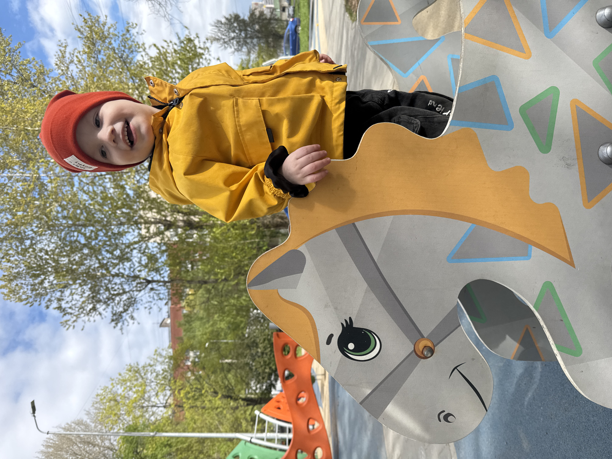 A young child wearing an orange beanie and a yellow jacket is standing on a gray and brown playground structure in the shape of a horse. The child is smiling and holding onto the horse structure. The playground is outdoors with trees and blue sky visible in the background. In the background, a building is visible. This playground is located in Saint Petersburg, Russia.