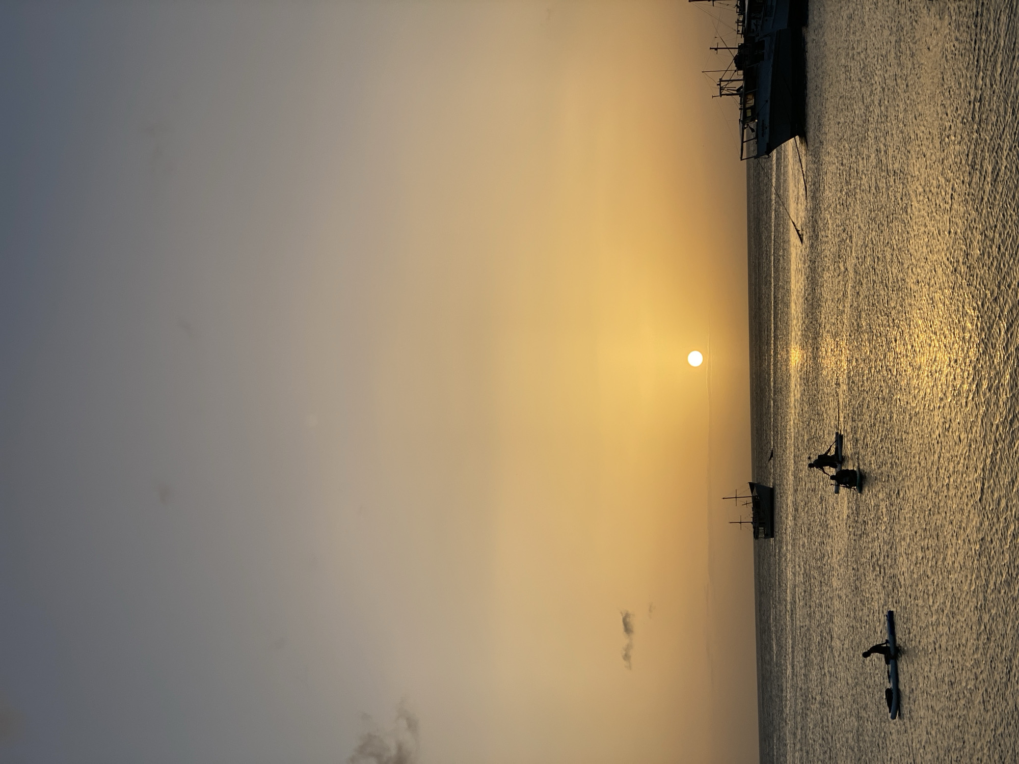An overcast, yet warm, sunset over the ocean at Juan Griego, Venezuela. The sky transitions from a pale blue to a soft orange as the sun sets. Several paddleboarders are visible on the calm water, their silhouettes stark against the reflection of the sun. In the distance, a few boats are anchored, adding to the serene coastal scene. The water is gently rippled, reflecting the warm hues of the sky.