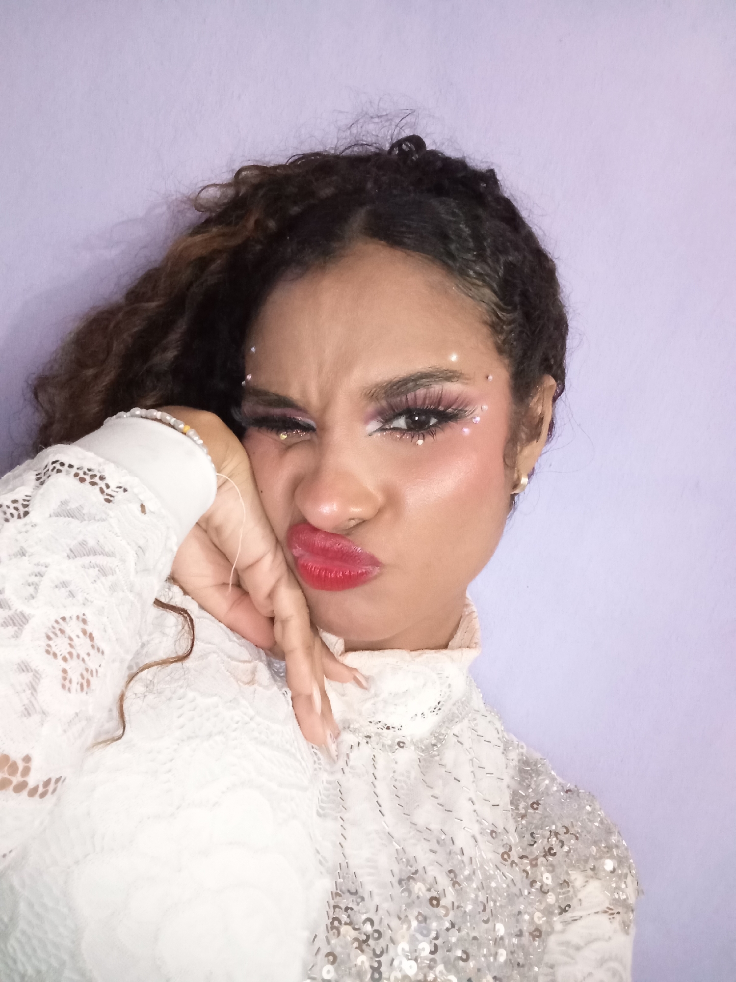 A young woman with curly brown hair is posing for a selfie indoors against a light purple wall. She is wearing a white, lace top with sequins on the shoulders and upper chest. Her makeup is elaborate, including false eyelashes, pink eyeshadow, rhinestones near her eyes, and bright red lipstick. She is making a pouting expression with her lips and resting her cheek on her hand, which has long, manicured nails. A pearl bracelet adorns her wrist. The location, Juan Griego, Venezuela, cannot be confirmed from the visual information provided.
