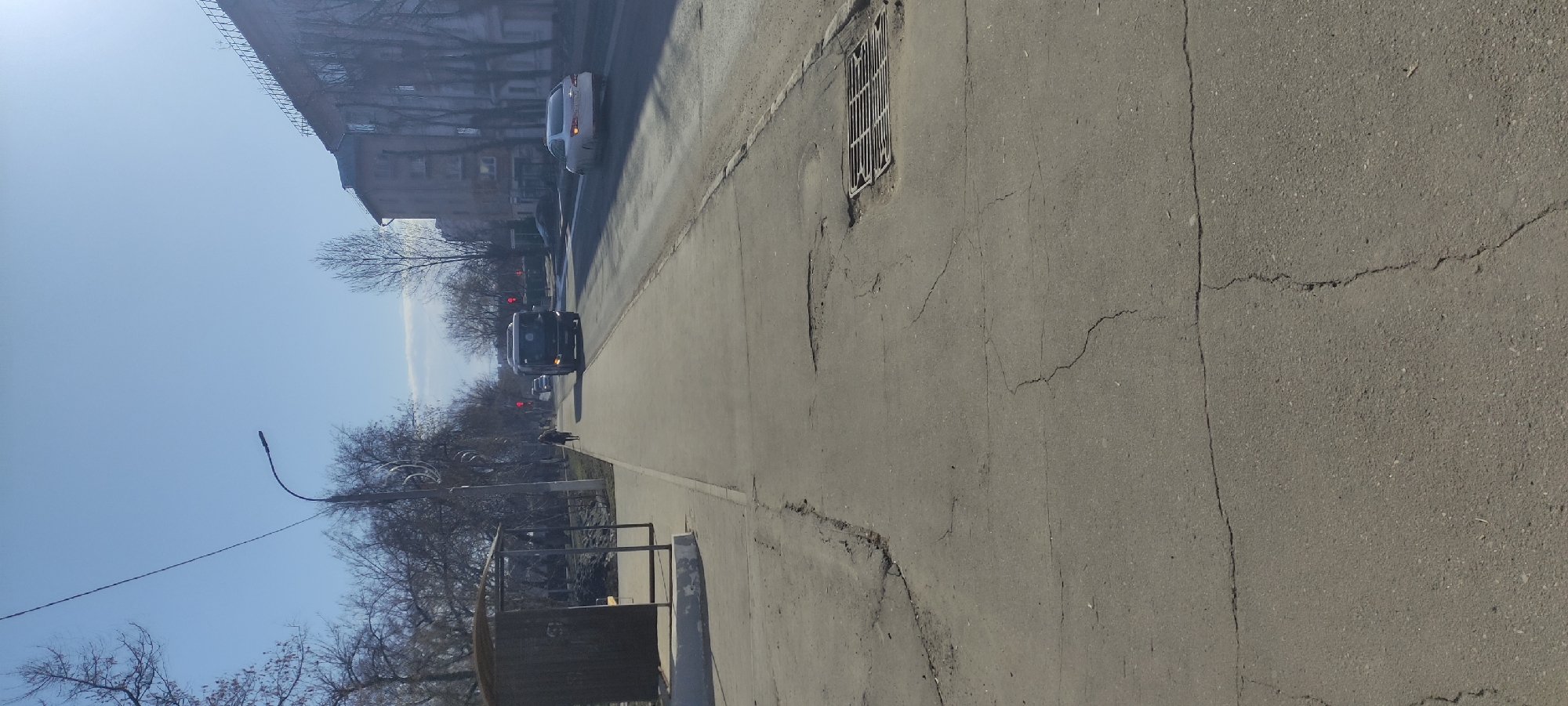 The image shows a street in Fryazino, Russia. The street has cracked asphalt and a manhole cover is visible. In the background, there are buildings and trees. Two cars are driving on the street. There is a bus stop to the right of the street. The sky is blue.