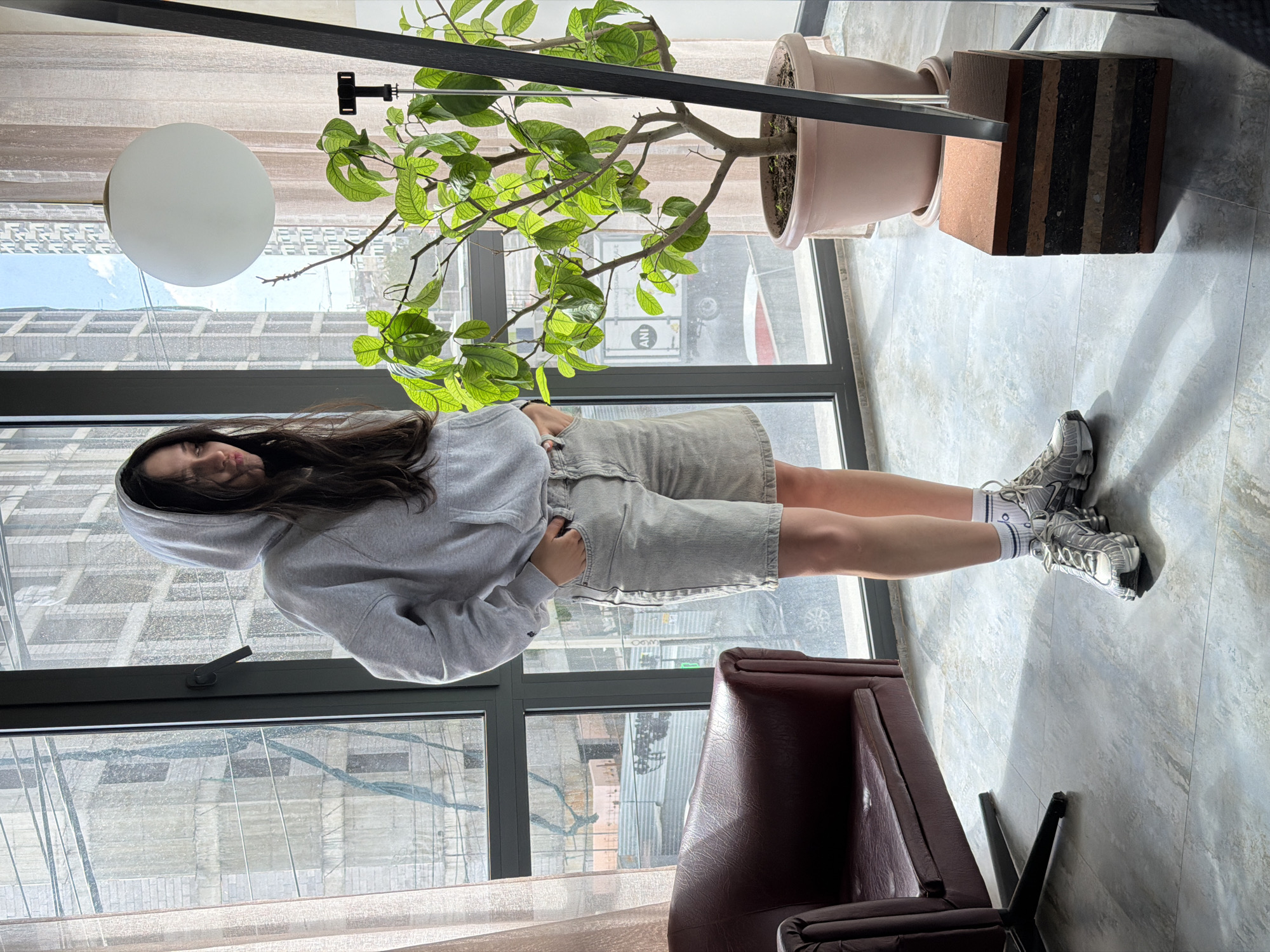 A medium, eye-level shot depicts a woman standing indoors near a window, against an urban backdrop, likely in Yerevan, Armenia. The woman is facing the left of the frame, sporting a light gray hooded sweatshirt and matching shorts, paired with white socks and gray athletic shoes. Her dark hair is loose, framing her face, and the hood is pulled up.
The environment includes a large window with a black frame, offering a view of city buildings. A brown leather chair is situated in the lower-right corner. A potted plant is positioned above, its leaves hanging down into the frame. A round, white pendant light is also visible. The floor appears to be tiled with a subtle marble pattern.