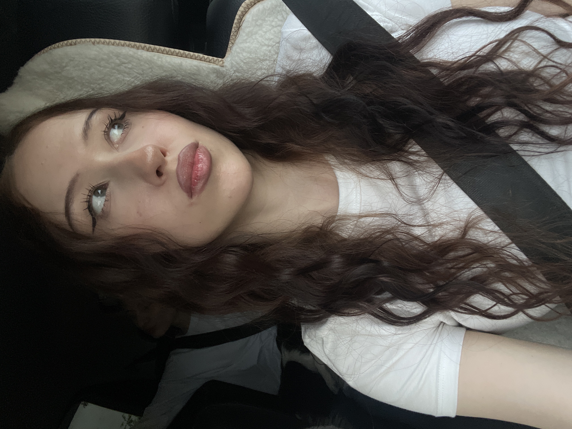 The media file is a close-up, vertically oriented, indoor photograph of a young person with long, dark brown, wavy hair. They are lying down, likely in a car, as indicated by the presence of a car seat headrest and a seatbelt. The person is wearing a white t-shirt. Their face is partially visible, with their eyes looking upwards. They have on makeup, including eyeliner and lip gloss. The lighting suggests it could be daytime, but the specific time of day and weather conditions are not discernible. The setting appears to be a private vehicle, possibly on a journey. No visible text is present in the image. The overall mood of the image is candid and relaxed.