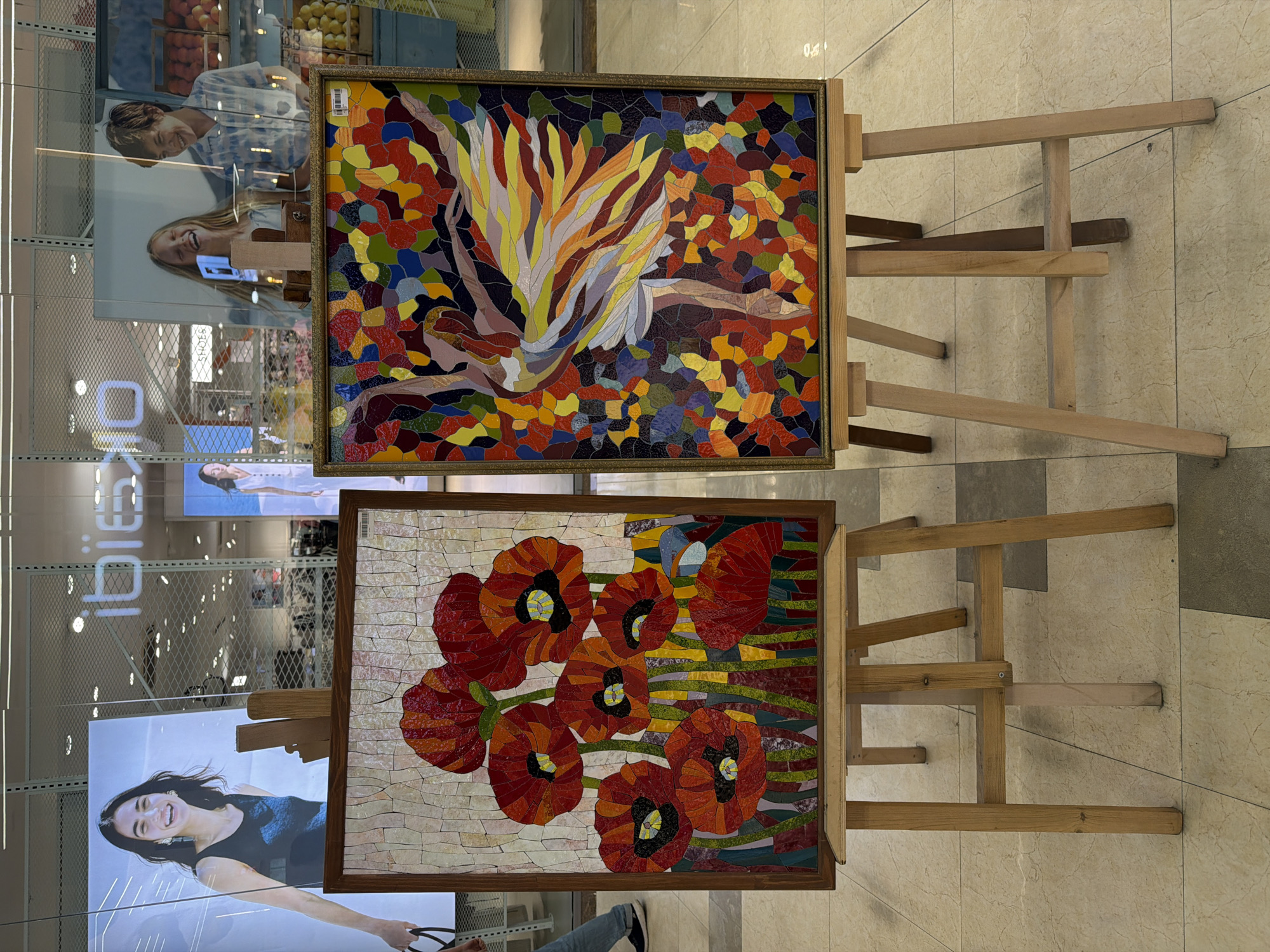 The image shows two framed mosaic artworks displayed on wooden easels. The top artwork features a bird-of-paradise motif in a mosaic of various colors set against a background of mosaic pieces in darker, more muted shades. The lower artwork is a depiction of red poppies on a background of pale mosaic pieces with green and yellow wavy stripes. Both artworks are on wooden easels. The background suggests an indoor location, possibly a gallery or shop, with reflective glass panels displaying images of people. The floor appears to be tiled with large, light-colored tiles. The location could be Yerevan, Armenia, if the person providing the request is located there. However, the image does not provide any information to confirm the location.