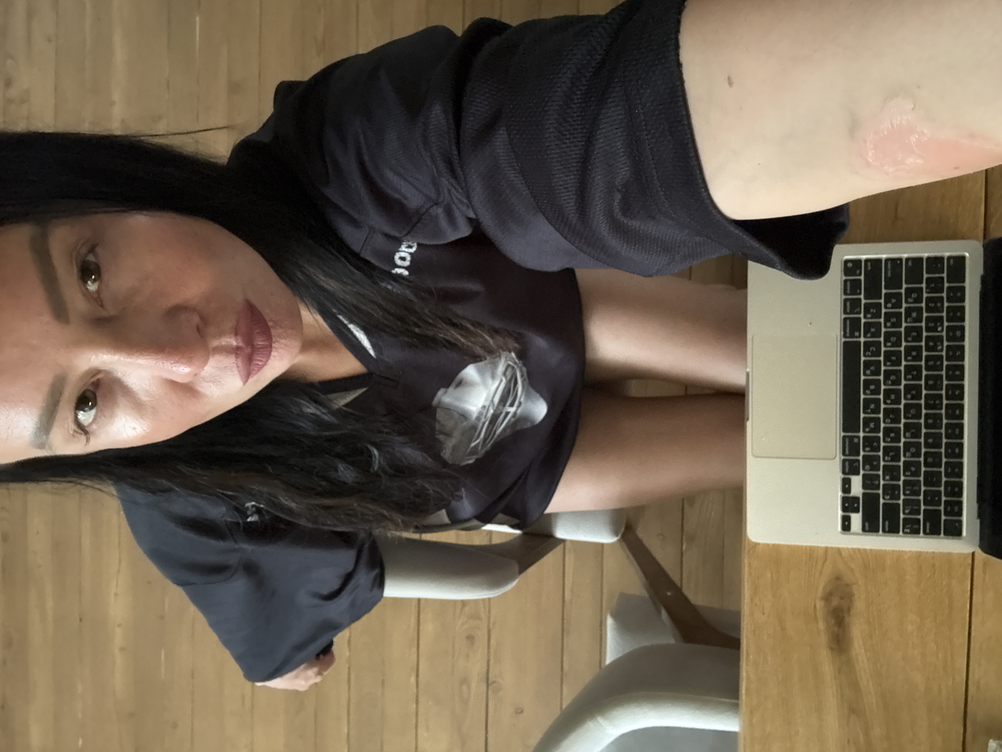 A woman with long black hair, dark lipstick, and fair skin is seated at a desk with a laptop. She is wearing a black shirt with a logo. A red mark is visible on her right arm. The desk has a wooden surface, and the floor is also wooden. Two chairs are visible. The location is Chaikovsky, Russia.