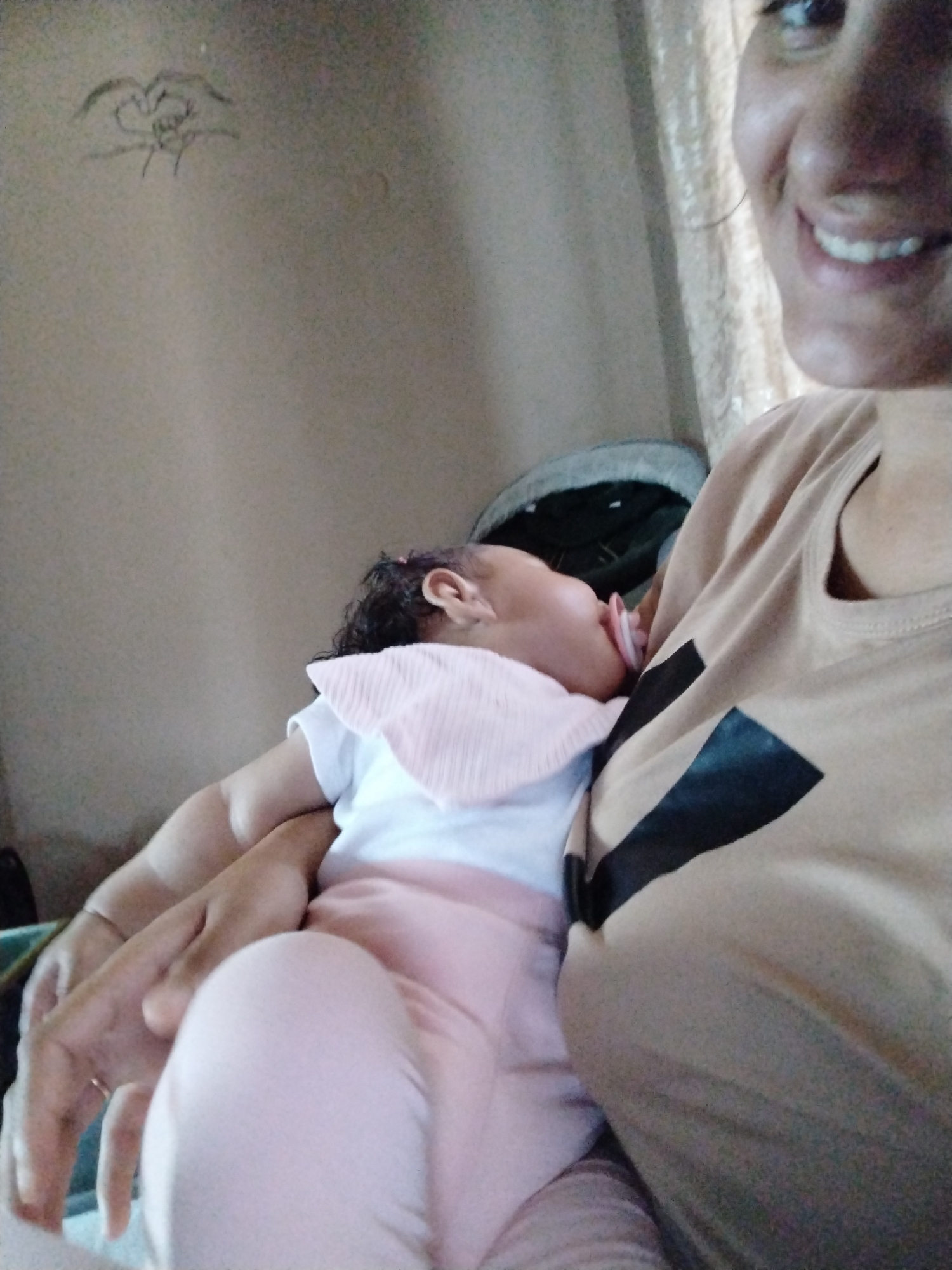 An indoor scene depicts a woman holding a baby in the city of Unknown, Unknown. The upper right portion of an adult female's smiling face is visible on the right, showing her right eye, part of her nose, and her mouth with visible teeth. She wears a light brown t-shirt featuring a black geometric design on the chest. A baby with dark, curly hair is positioned on the left, held by the woman, and appears to be asleep with a pink pacifier in its mouth. The baby is dressed in a white top, a pink ribbed bib, and light pink pants. The woman's hands are visible supporting the baby, one wrist bearing a thin bracelet. The background consists of a plain light beige wall, with a rough sketch of two hands forming a heart shape visible in the upper left corner. A light-colored, possibly textured curtain is partially visible on the far right behind the woman's head, and a dark object, possibly part of an infant carrier or stroller, is partially visible behind the baby on the lower left.
