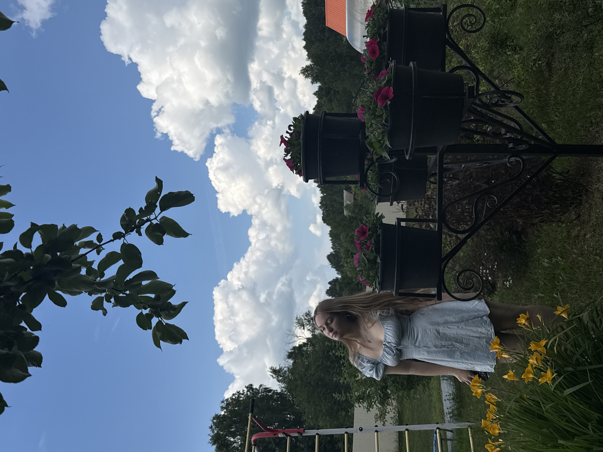 A young woman with long blonde hair stands outdoors in a garden in Zhigalki, Russia, wearing a light blue, puff-sleeved dress. She is positioned to the left of a black metal multi-tiered plant stand filled with flowering plants, some of which have pink petals. In the foreground, a patch of yellow lilies blooms. Behind the woman, a white fence and a children's playground structure are visible, along with trees and the roof of a building in the distance. The sky above is blue with large, fluffy white clouds, and tree branches with green leaves extend into the upper portion of the frame. The lighting suggests it is daytime, possibly late afternoon, with the sun casting soft shadows.