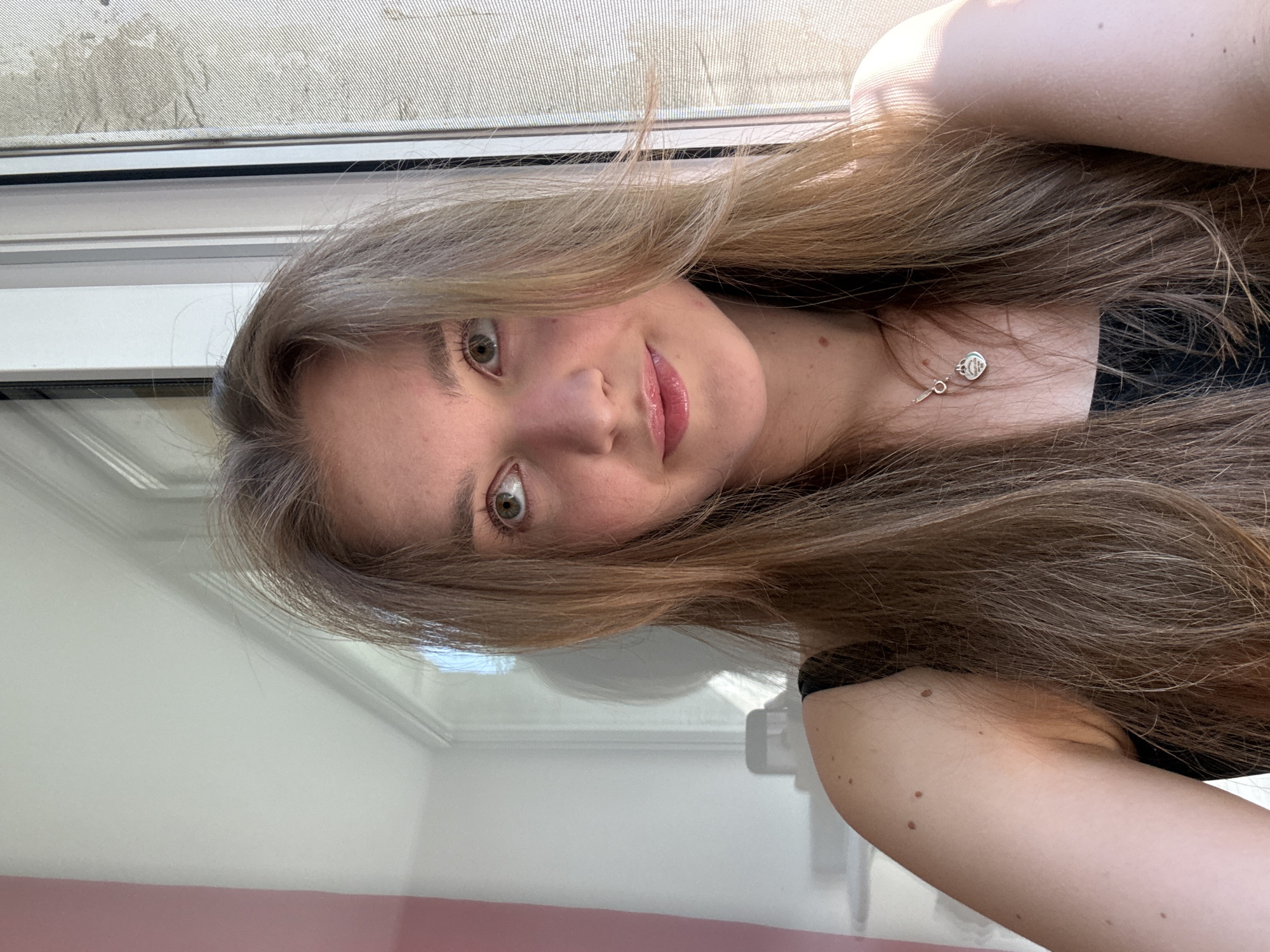 A young woman with long, light brown hair is posing for a selfie indoors. She has light skin, green eyes, and a necklace with a heart-shaped pendant. She wears a black top. The background features a window with a screen and a white frame. Part of a light pink wall is visible. The image was likely taken in a private residence. There's no concrete indication that the image was taken in Perm, Russia.