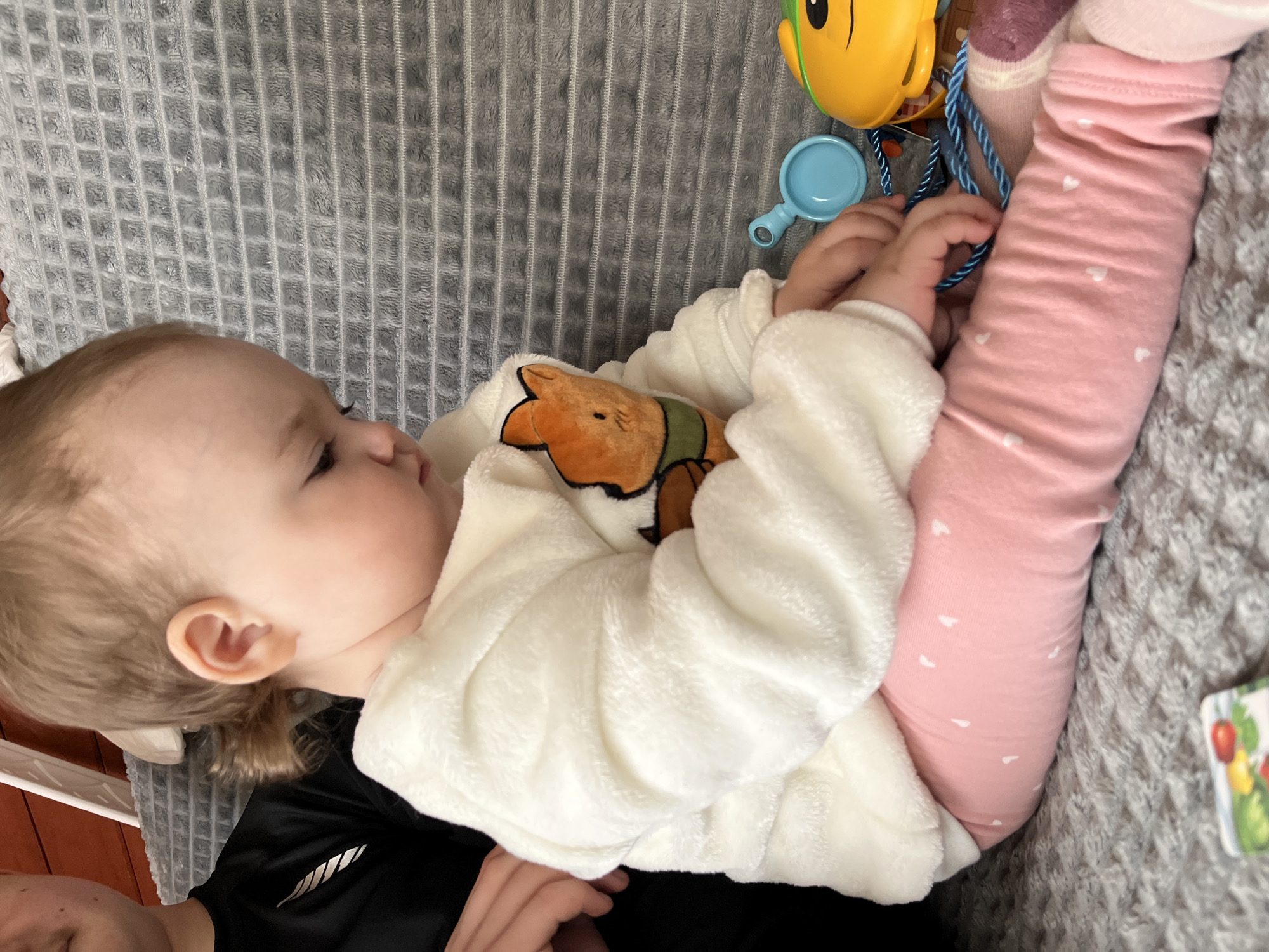 A child, possibly a toddler, is lying on a gray blanket or textured surface. They are wearing pink leggings with small white heart patterns and a white, plush jacket with an orange and green cartoon bear design. The child's left arm is partially visible, and their hands appear to be holding a blue string or cord attached to a yellow toy. Also nearby is a small blue toy resembling a magnifying glass. A dark-colored shirt or clothing is partially visible beneath the child's jacket, suggesting the presence of another person holding the child. A book with what appears to be vegetables printed on the cover, is partially visible at the bottom right of the picture. The lighting is soft and diffused, and the overall setting appears to be indoors. There is no information to suggest that the location is in the city of Мшинская, Russia.
