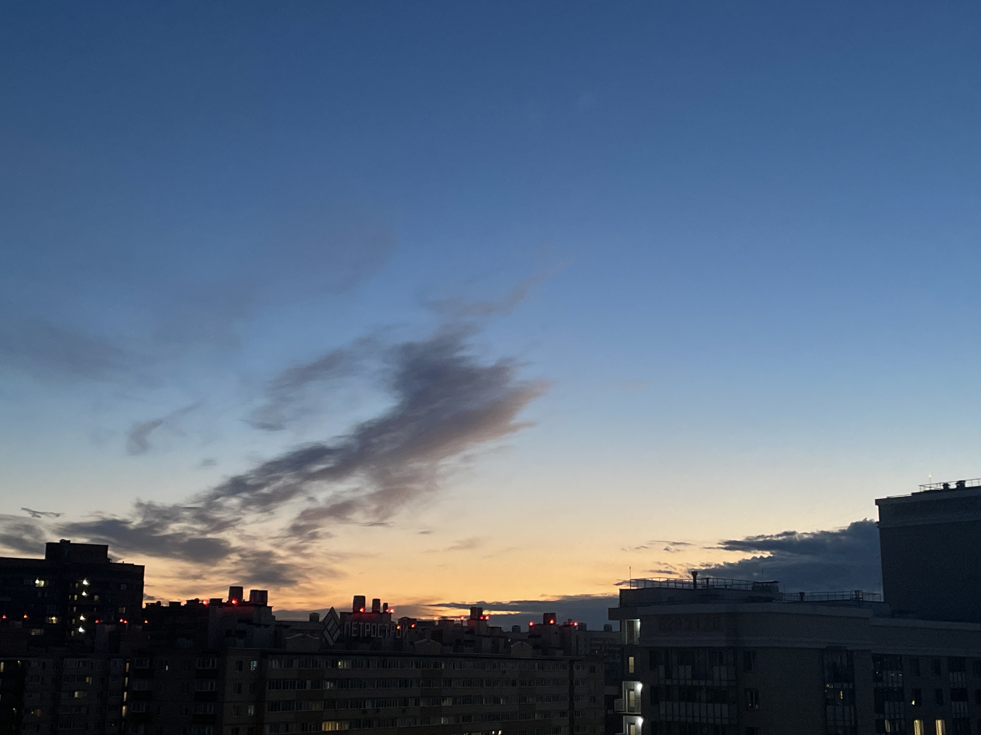 An exterior, eye-level, medium shot captures a dusky sky above a cityscape in an unknown location in Russia. The sky is a gradient of deep blue at the top, transitioning to a pale orange and yellow near the horizon, with wispy grey clouds scattered throughout. Silhouetted against the sky are several buildings of varying heights. Some buildings have illuminated windows, indicating an urban environment at dusk or night. Several buildings feature red lights on their rooftops. The image is a static composition, with no discernible actions or interactions taking place. The overall impression is one of a quiet urban evening.
