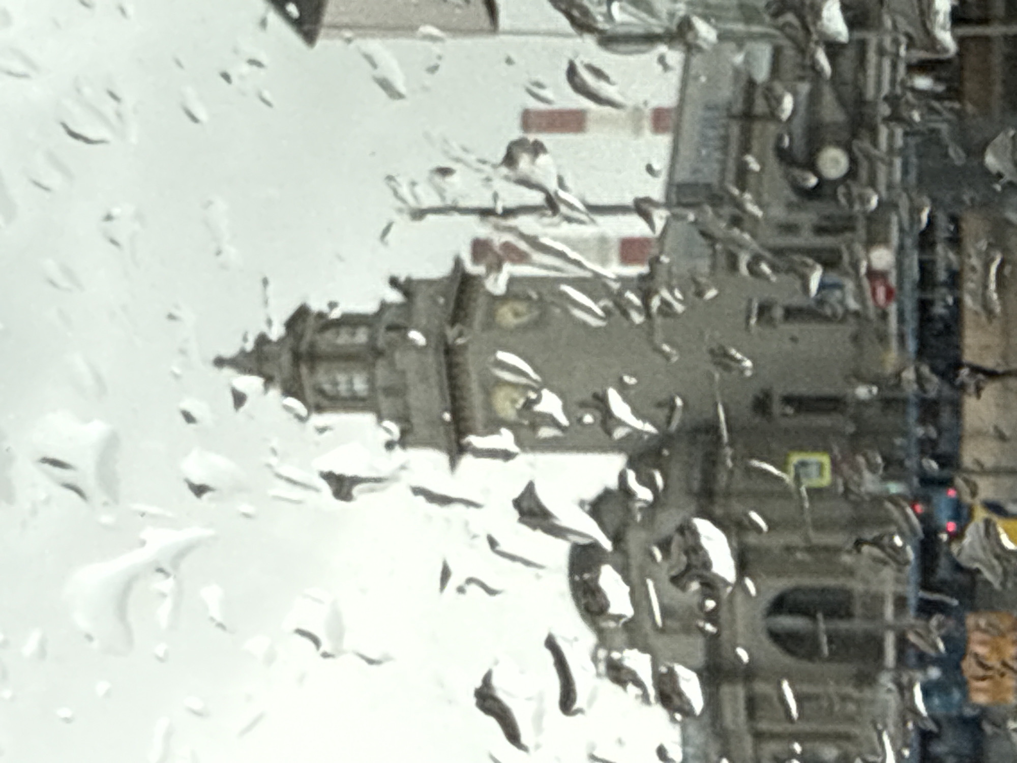 The image shows a cityscape through a rain-covered window. A building with a tower is visible to the left. To the right, there are other buildings and possibly a street scene with a vehicle and pedestrians.
Based on the building architecture, the scene is likely in Moscow, Russia.