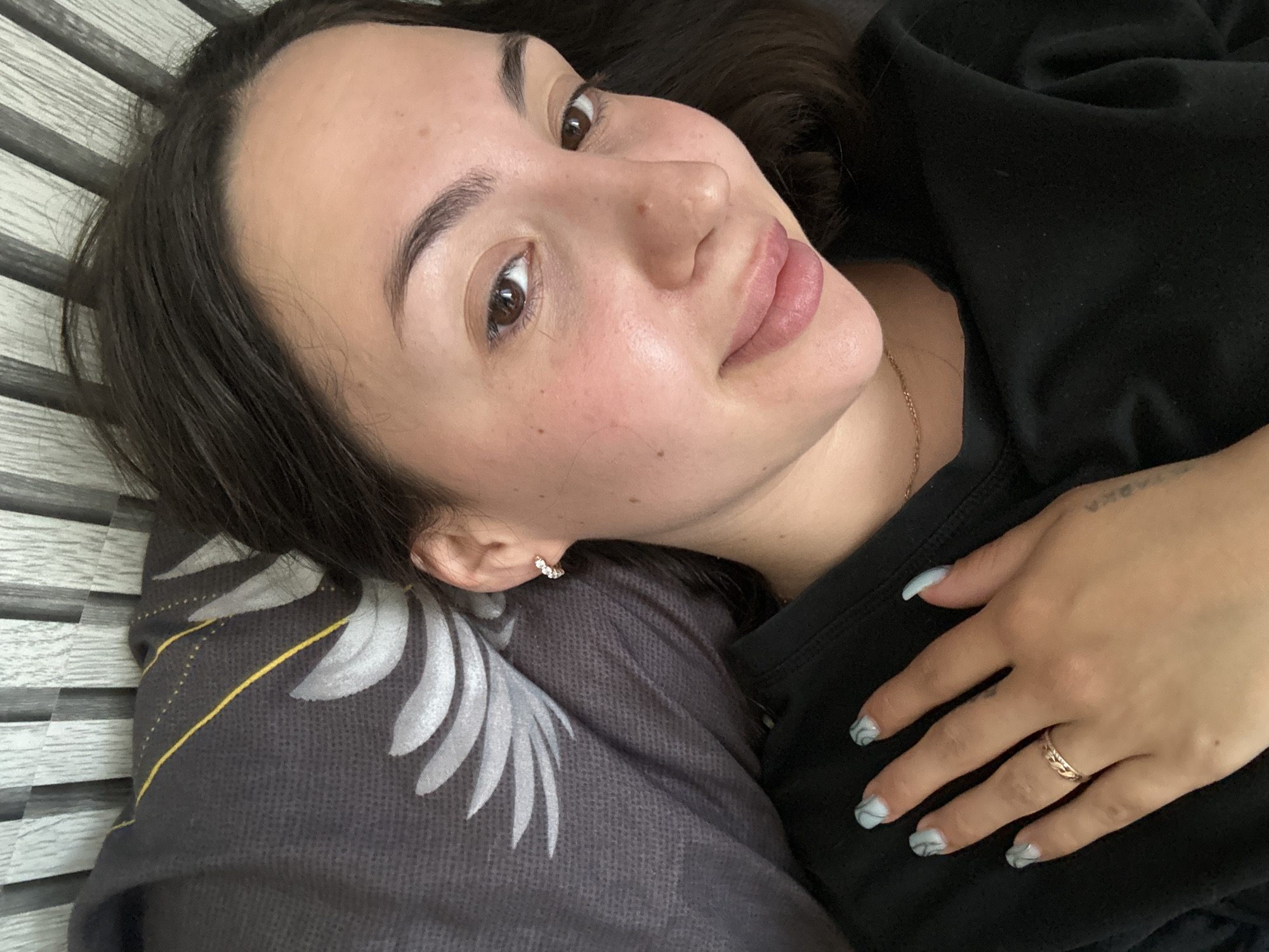 The image shows a close-up, high-angle selfie of a young woman lying in bed. She has dark hair, brown eyes, and fair skin with some light redness on her cheeks. She is wearing a black t-shirt and a thin gold necklace. Her left hand rests on her chest, revealing light blue marble-patterned fingernails and a rose gold ring on her ring finger. A decorative pillow with a gray background and a white feather pattern is visible behind her head. The background behind her hair appears to be a textured wall with vertical slats. The image is taken indoors, likely in a bedroom. No specific location in Марковский, Russia can be determined from the image.