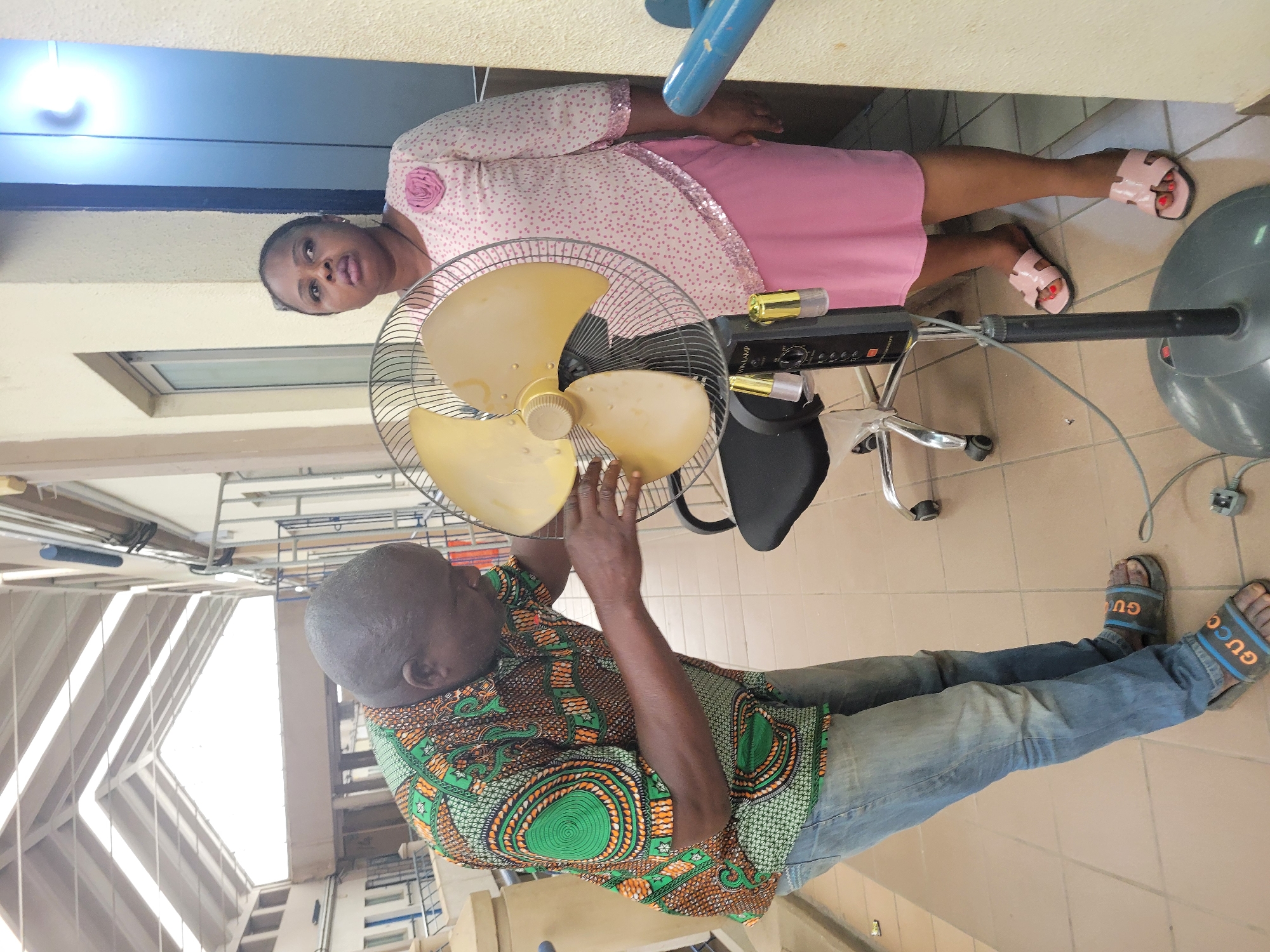 A man wearing a patterned shirt and jeans appears to be repairing a yellow standing fan. A woman wearing a pink dress stands next to the fan. The location is likely in Abuja, Nigeria. The environment is an indoor space, possibly an office. The floor is tiled. The man's hands are touching the fan blades. Both individuals are looking towards the camera. The man is also wearing blue Gucci slippers. The woman wears pink sandals with red nail polish.
