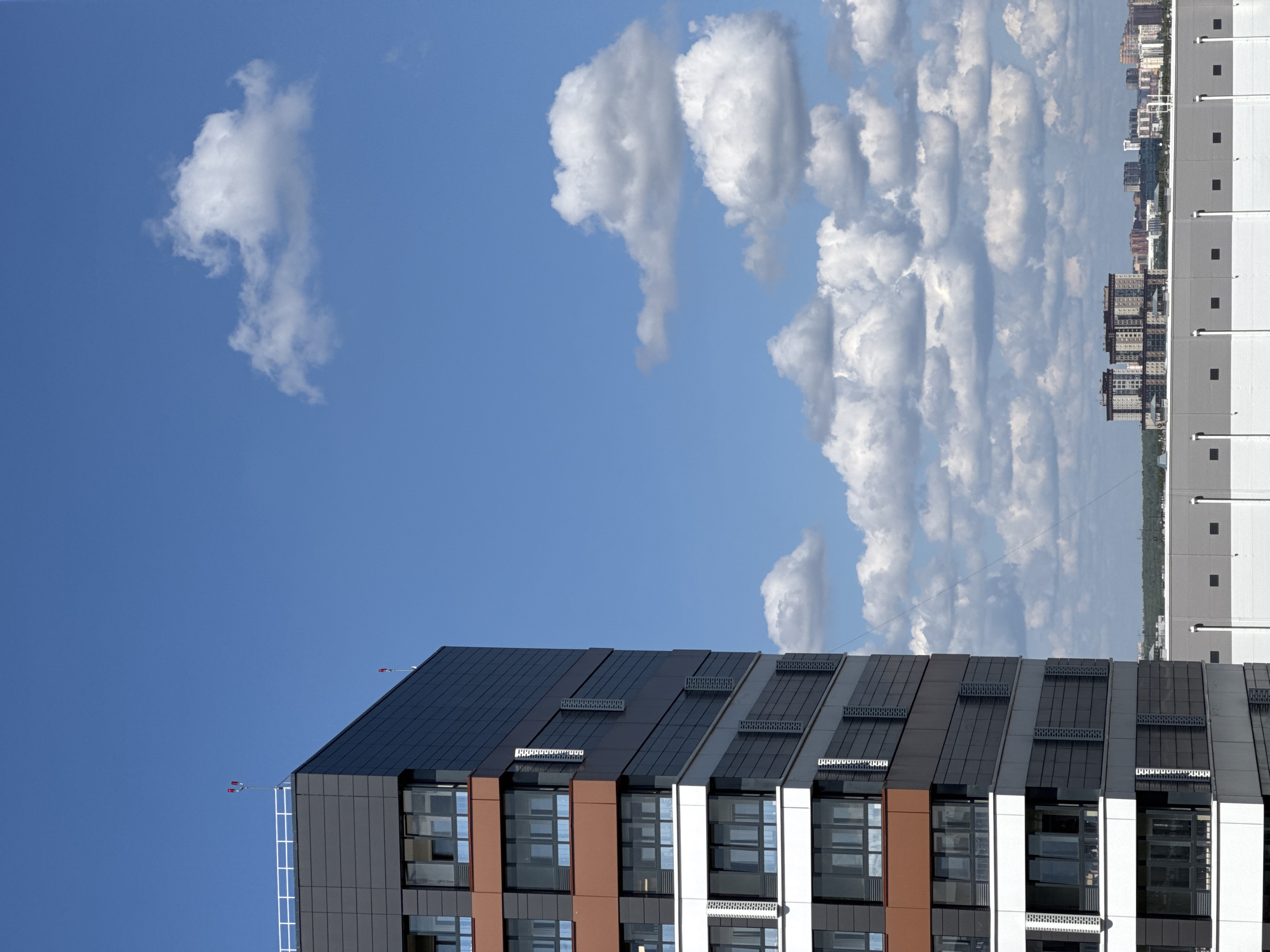 The image depicts a modern, multi-story building with a dark gray facade and orange accents, set against a bright blue sky with scattered white clouds. The building features numerous windows and architectural details. In the background, a distant cityscape is visible, characterized by a cluster of buildings with varied architectural styles, suggesting an urban environment. A large, low-lying structure with a gray and white facade is also present in the foreground, partially obscuring the distant city. The scene appears to be captured on a clear, sunny day. The provided context places the image in Novosibirsk, Russia.