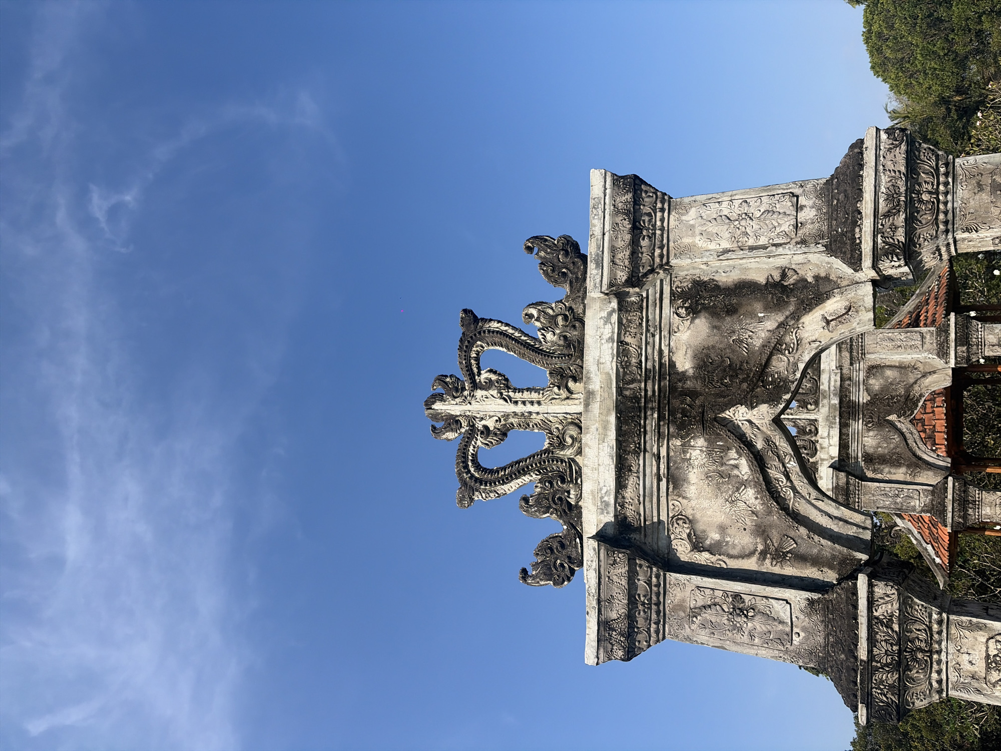 The image displays a detailed, ornate stone archway topped with intricate carvings, possibly depicting a crown or stylized dragon. The stone is weathered and shows signs of age and possibly moss or discoloration in some areas. The archway features detailed relief work on its pillars and across its lintel, with floral and avian motifs visible. Below the main arch, the roof of a smaller structure with terracotta tiles can be seen. The backdrop is a clear, vibrant blue sky with thin, wispy clouds. Lush green trees frame the lower right portion of the image, suggesting an outdoor setting. A tiny, pinkish dot, possibly a kite or a distant object, is visible in the sky to the left of the arch's apex. The scene is likely located in Karangasem, Indonesia, given the architectural style which is characteristic of Balinese temples or historical structures. No people are visible in the frame.