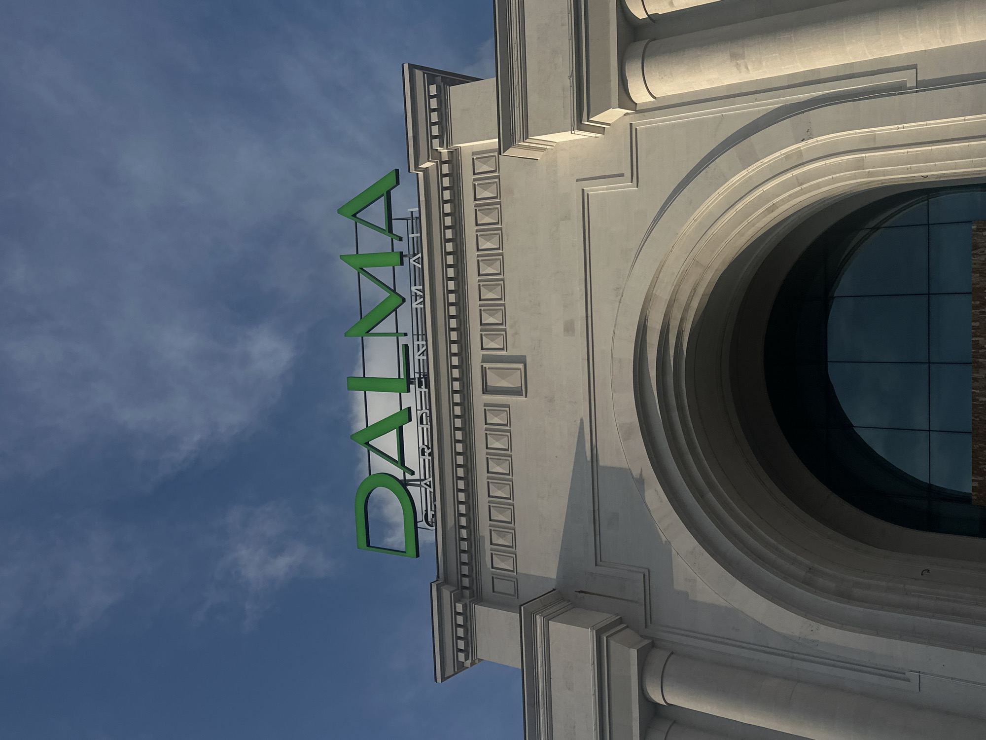 The image shows the exterior of the Dalma Garden Mall, a shopping center located in Yerevan, Armenia. A large arched window is visible on the right side of the frame. Above the window, the name "DALMA GARDEN MALL" is displayed in large, green lettering against a blue, cloudy sky. The architectural style appears to be modern with classical elements.