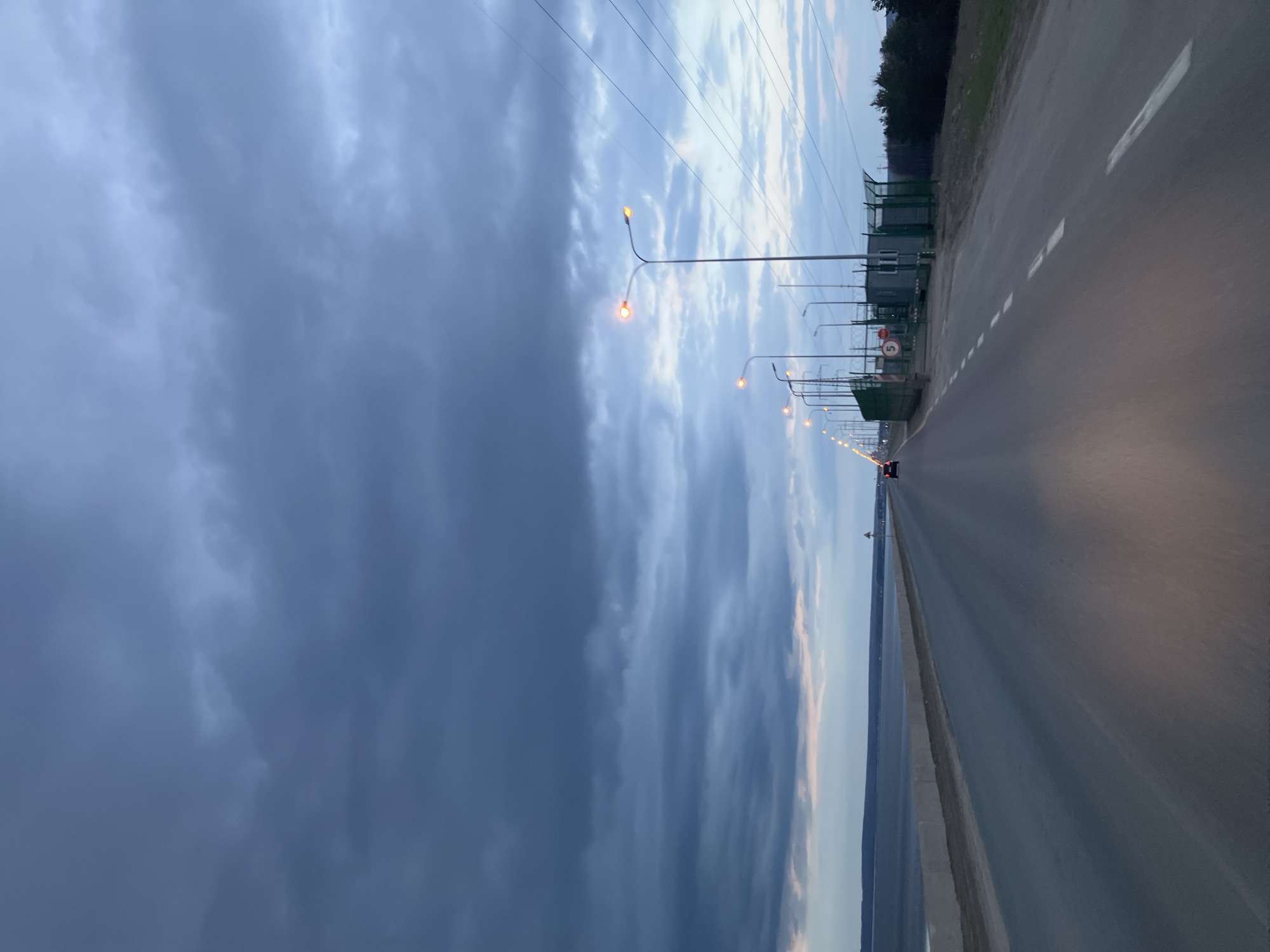 The image, taken in Noviy, Russia, captures a highway or causeway stretching into the distance alongside a vast body of water under a dramatic sky. The road features white dashed lane markings, and a series of streetlights with orange-yellow illumination line its left side, indicating dusk or early night. Overhead, multiple power lines traverse the frame against the cloudy backdrop.
To the left of the road, a fenced-off area contains utilitarian buildings behind green mesh fencing, suggesting an industrial or utility facility. A red circular sign, likely a speed limit, with the number "5" in white, is clearly visible attached to the fence, along with an orange and white lifebuoy. Further along the road, the red taillights of a distant vehicle are discernible. A dark tree line marks the horizon beyond the infrastructure on the left. The sky is heavily overcast with varying shades of grey clouds, though some lighter areas are also present. A small, indistinct object, possibly a sailboat or a navigational marker, can be seen on the distant water.
