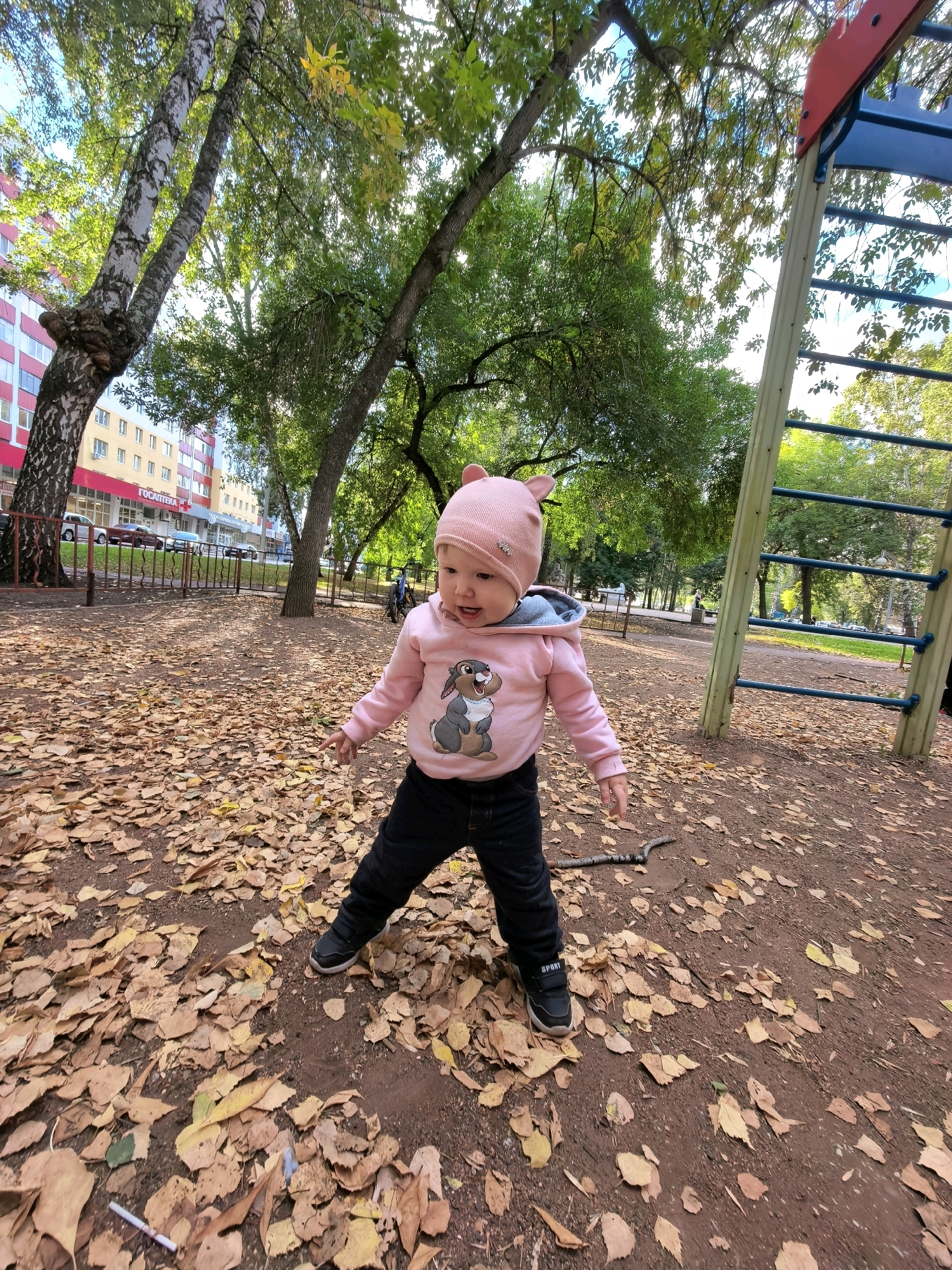 In Sterlitamak, Russia, on an autumn day, a young child wearing a pink hooded sweatshirt with a bunny graphic and a matching hat with ear accents stands on a playground covered in fallen leaves. The child is wearing black pants and sneakers. To the right, a section of a playground structure with wooden supports and blue horizontal bars is visible. Behind the child, trees with sparse autumn foliage stand, and in the background, a multi-story building with red and beige facades can be seen, along with parked cars on a street. The overall scene depicts a child playing outdoors in a park setting during the fall season.