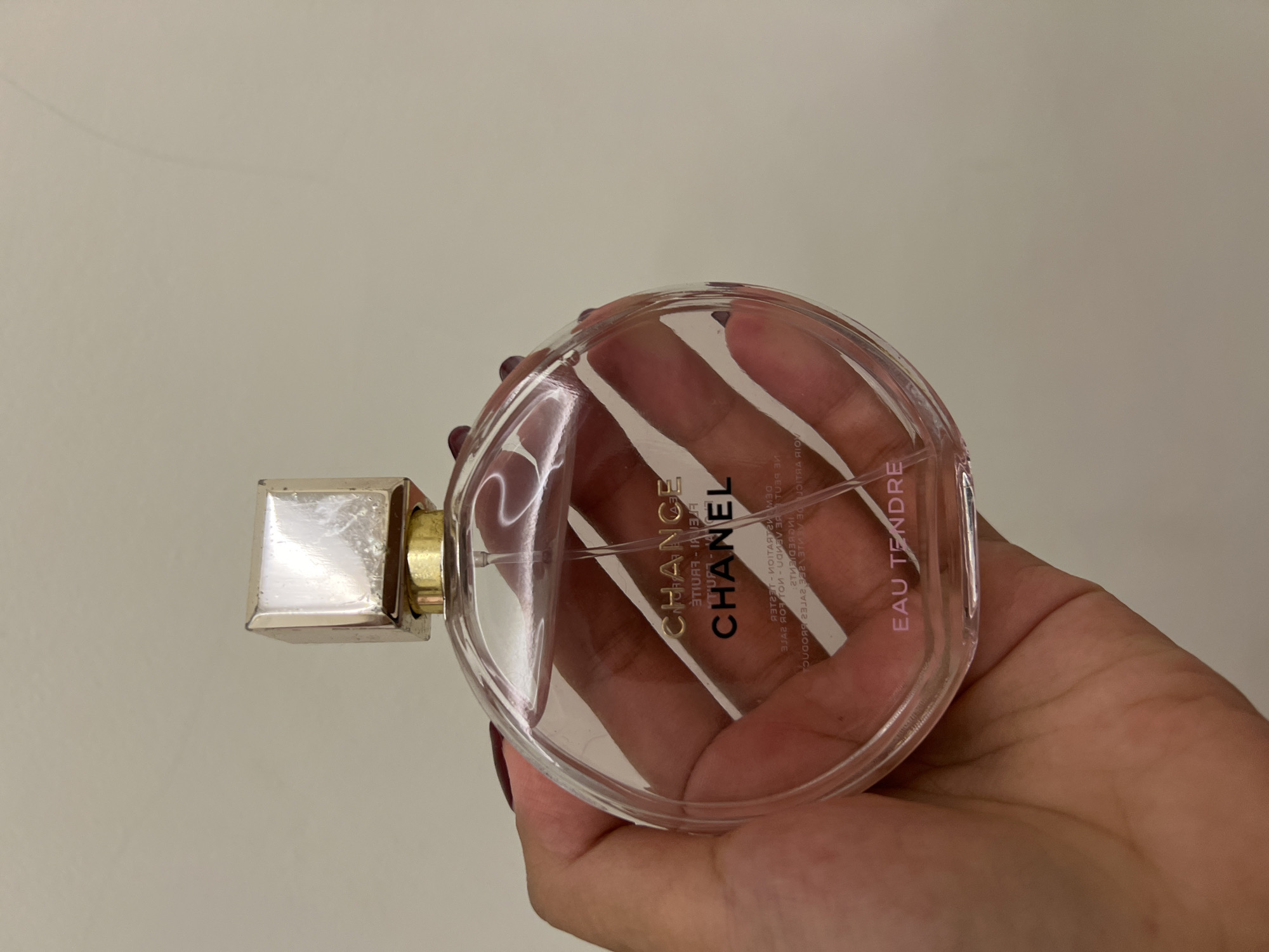 The image displays a close-up, vertical shot of a human hand holding a transparent, circular perfume bottle against a plain, light-colored background.
The subject is a hand, likely female, grasping the bottle from the underside and sides. The fingernails are painted with a dark red or burgundy polish. The fingers are visible through the clear glass of the bottle.
The primary object is a "CHANEL CHANCE EAU TENDRE" perfume bottle. It is round and flattened, made of clear glass, and contains a light pink liquid. A transparent dip tube is visible extending from the sprayer mechanism to the bottom of the bottle. The front of the bottle clearly displays "CHANCE" in gold lettering, "CHANEL" in black lettering, and "EAU TENDRE" in light pink lettering towards the bottom. Reflected or printed on the reverse side and visible through the liquid and front text, are several lines of smaller, mirrored text including "FLEUR FRUITÉ", "ÉDITION LIMITÉE", "TESTER - NE PEUT ÊTRE VENDU" (Not for Sale), and "VOIR ARTICLE" (See Item). The bottle is topped with a square, metallic cap that appears silver-toned but shows signs of wear, scratches, and slight tarnish. The cap sits on a narrow gold-colored collar.
The immediate environment consists of a plain, light-colored background, exhibiting no features that would indicate a specific geographical location, such as the city of Mengwi, Indonesia, or any other specific locale. The lighting is soft and even, suggesting an indoor setting.