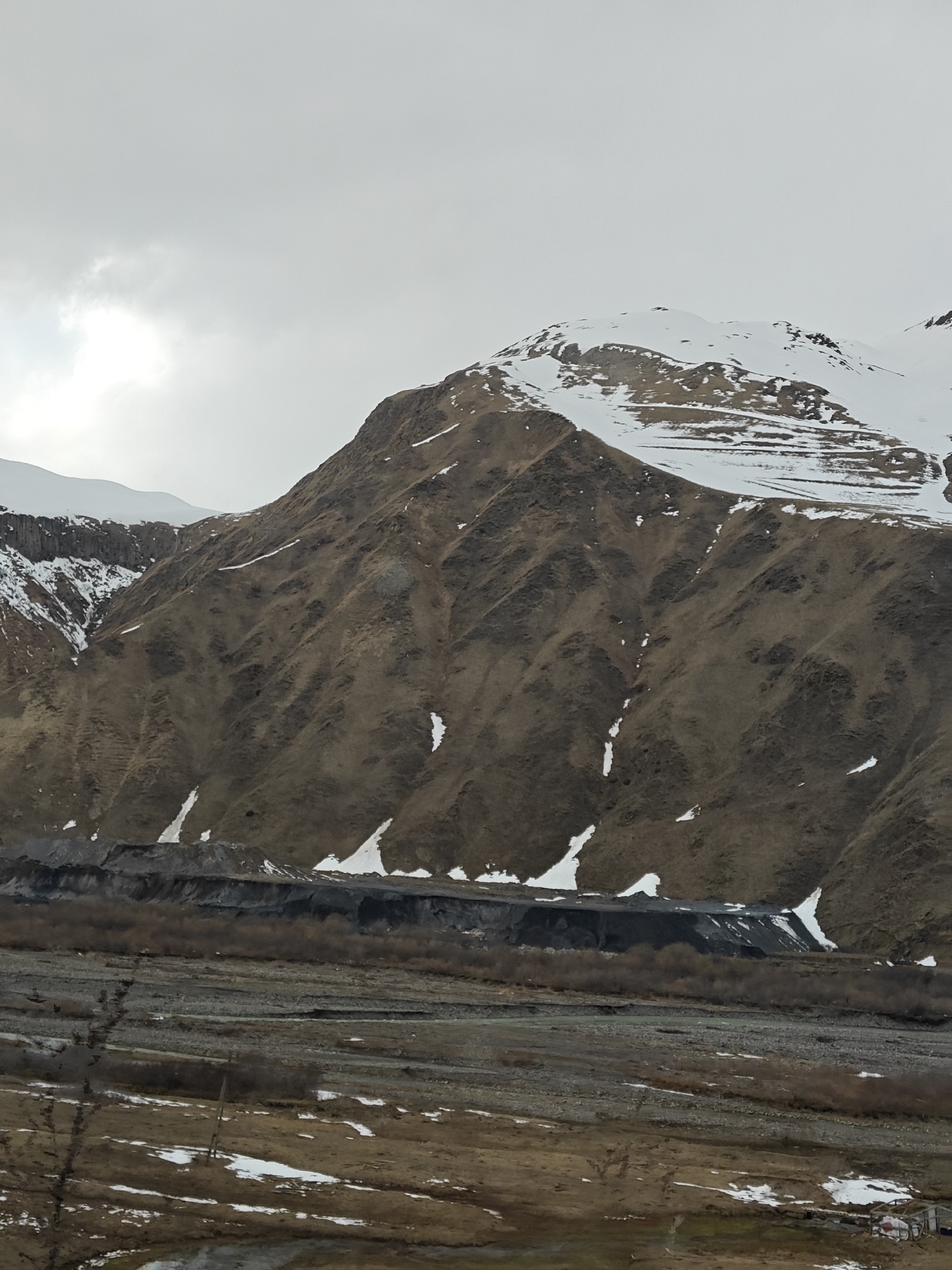 The image captures a vast, rugged mountain landscape under a uniformly overcast, light grey sky. In the foreground, a wide, rocky riverbed, appearing mostly dry or with minimal water, stretches horizontally across the frame. The ground immediately beyond the riverbed is a mix of brown, sparse vegetation and melting snow patches.
The dominant features are the large mountains in the midground and background. Their slopes are primarily covered in dry, brown vegetation or bare earth, suggesting a cold season like late winter or early spring. Patches of white snow are scattered across these slopes, particularly in depressions and gullies. The highest peaks, especially towards the right side of the frame, are capped with significant amounts of bright white snow.
At the base of the main mountain range, just above the riverbed, there is a distinct, darker band of disturbed ground, possibly indicating a landslide, erosion, or a man-made alteration, which stands out against the lighter brown slopes above it. The diffused lighting from the cloudy sky creates a somewhat stark and muted palette. No people, animals, or specific activities are depicted, giving the scene a sense of immense scale and solitude. There is no visible text in the image.