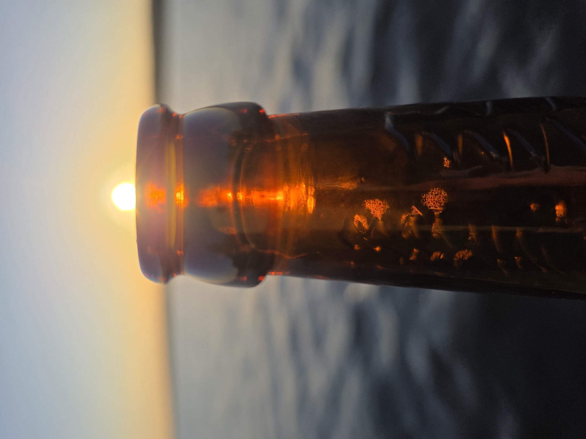 The image shows the neck of a brown glass bottle held up to the setting sun. The sun appears as a bright orb at the opening of the bottle, casting a warm glow on the glass. The background is a blurred landscape of water and sky, suggesting a waterfront location. The scene is likely in Chaikovsky, Russia, as specified in the prompt. No people are visible in the image. The bottle is the primary object, with the sun and landscape serving as the backdrop. The action is the deliberate positioning of the bottle to capture the sun within its opening. The interaction is between the bottle, the sun, and the environment, creating a visually interesting effect.