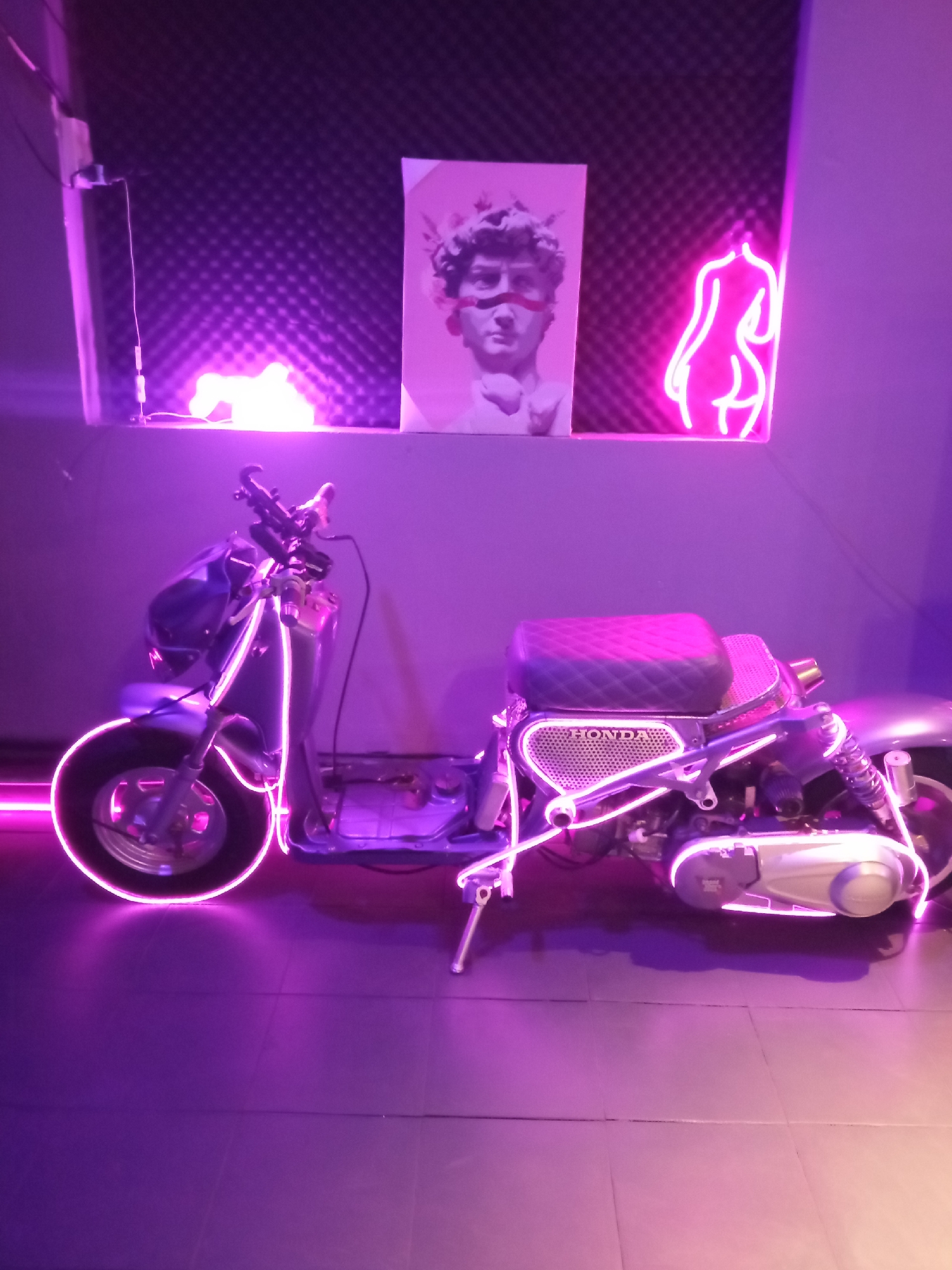 The image shows a scooter adorned with pink neon lights, parked indoors in what appears to be a dimly lit space with purple ambient lighting. The scooter is silver and purple, with a diamond-patterned seat. The "HONDA" logo is visible on the scooter's side.
In the background, there is a textured dark wall with a framed print of a classical sculpture's head, possibly David, with red accents on its face. To the right of the print, a pink neon outline of a human figure is displayed. In the foreground, the floor is tiled and also reflects the purple lighting. The overall atmosphere is artistic and modern, with the neon lights creating a vibrant, futuristic aesthetic. This scene does not provide any contextual information or visual cues to suggest it is located in Nha Trang, Vietnam.