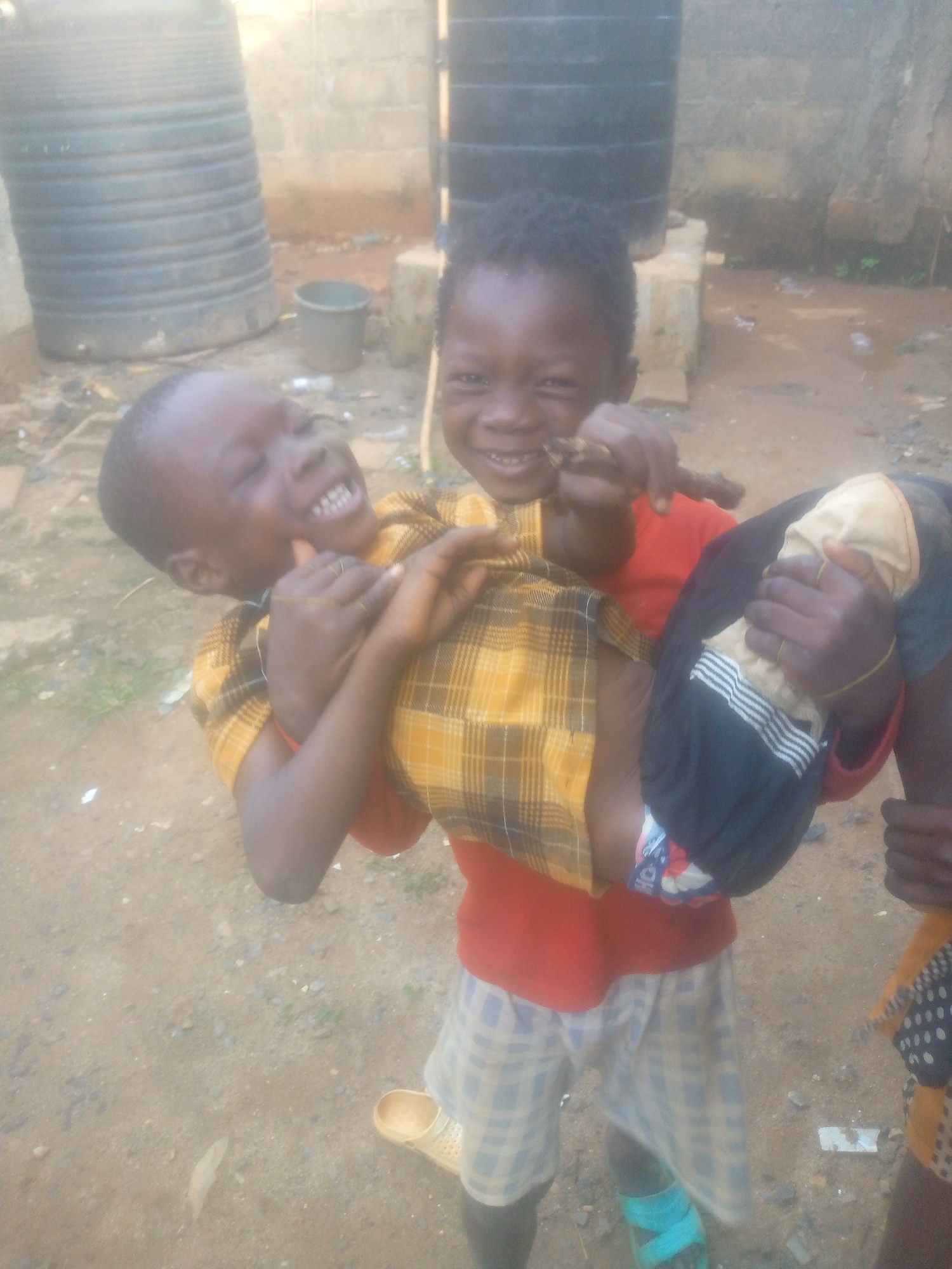 Two young children are captured mid-play in an outdoor setting, both displaying wide, joyful smiles. The child on the right, dressed in a red t-shirt and light blue and white plaid shorts, is carrying the other child on his hip. He wears a turquoise sandal on his left foot and holds a small, brown, stick-like object in his right hand. The child being carried is laughing heartily, with his head tilted back and mouth open, revealing his teeth. He is wearing a yellow and black plaid shirt over what appears to be a red garment, and dark shorts with a white striped hem.
The background features a rustic environment with a dirt ground scattered with small pebbles and sparse debris. To the left and upper right, two large, dark-colored water tanks, possibly barrels or drums, are visible. A small, dark bucket sits on the ground between them, and a concrete block wall can be seen behind the tanks. The lighting indicates it is daytime, likely a bright but possibly overcast day, as there are no harsh shadows. The overall scene conveys a sense of innocent playfulness and happiness between the two children. No visible text is present in the image.