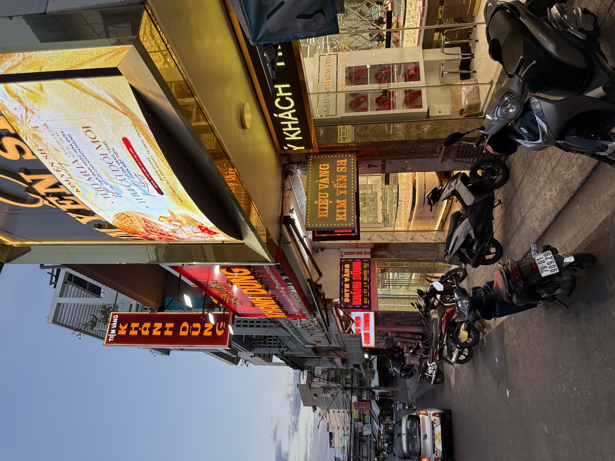 The image depicts a street scene in Nha Trang, Vietnam, likely during the evening or early morning, given the lighting and the illuminated signs. The primary focus is on a row of businesses, predominantly jewelry stores, indicated by prominent signs such as "TIỆM VÀNG KHANH DUNG" and "HIỆU VÀNG KIM YẾN SH," which translate to "Gold Shop Khanh Dung" and "Gold Shop Kim Yen SH" respectively. These signs, illuminated with red and yellow neon lights, suggest a commercial district.
In the foreground, several motorcycles are parked along the sidewalk, typical of Vietnamese street life. One black motorcycle in the immediate foreground has a license plate reading "79-D1 909.29." Further down the street, a silver car is also visible.
The buildings lining the street are a mix of modern and older architecture. The jewelry stores feature large display windows showcasing what appear to be gold jewelry, illuminated from within. One store has a prominent digital billboard displaying advertisements. In the background, taller buildings are visible, suggesting an urban environment. The sky is a dusky blue with some cloud cover. The street itself is paved, and the sidewalks are a combination of concrete and stone. There are no distinct individuals prominently featured in the image, though silhouettes or distant figures might be present within the shops or on the street. The overall impression is one of a bustling commercial street in a Vietnamese city.