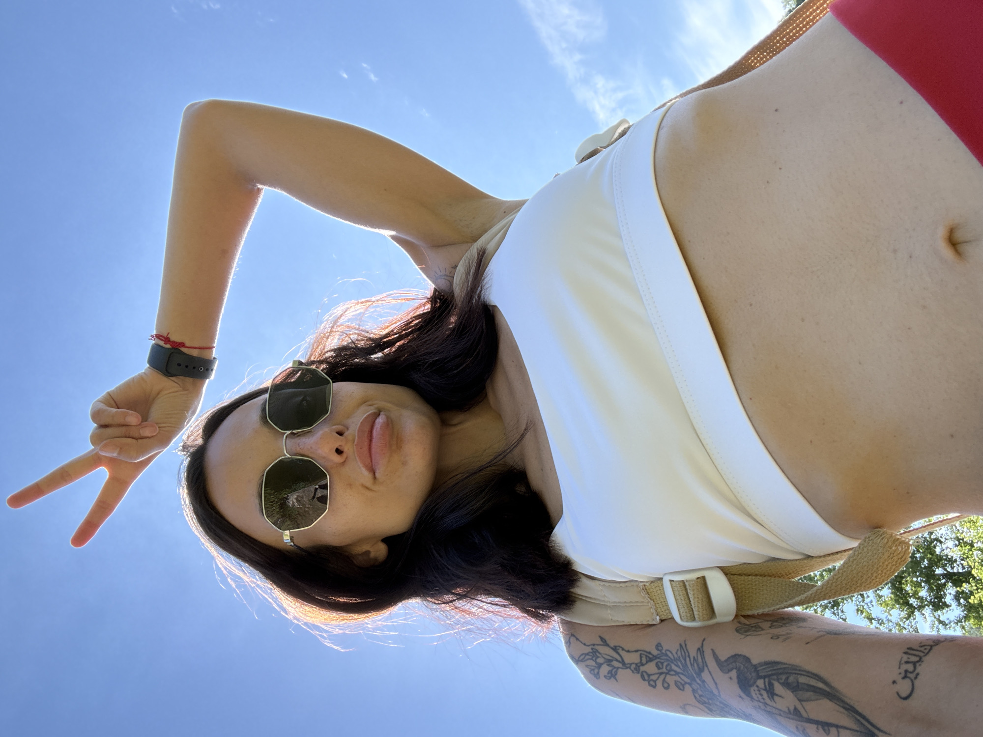 The image is a low-angle, outdoor selfie taken against a clear blue sky with some scattered clouds. A young woman with dark hair is in the foreground, wearing sunglasses, a white crop top, and red bottoms. She is raising her right arm, with her index and middle fingers extended in a peace sign. She has tattoos on her left arm and is wearing a watch on her right wrist. A light-colored backpack is visible over her shoulders. The background shows greenery, suggesting a park or natural setting, possibly within the city of Pleven, Bulgaria, given the prompt's context. The lighting indicates it is daytime, likely sunny.