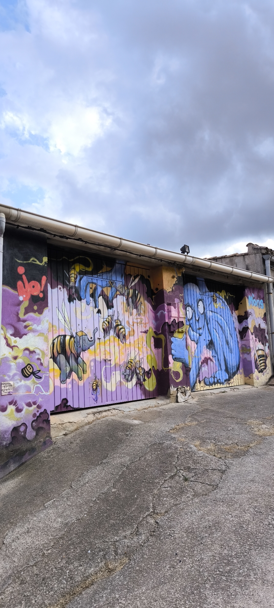 The image depicts a vibrant mural painted on a corrugated metal garage door in Fanzara, Spain. The artwork features cartoonish characters and a surreal scene involving bees. On the left, a red "Io!" is spray-painted. A bee-like elephant with striped wings and body is depicted in the center, surrounded by several bees in various sizes. To the right, a large, blue, cartoonish creature with wide eyes and a surprised expression is rendered, holding a yellow object. The word "Mons" is written in yellow letters to the right of the blue creature. The mural covers the entire width of the garage door, which is a light purple color. The surrounding environment shows a concrete ground with some cracks and dry grass, and a cloudy sky above. A white gutter runs along the top of the garage.