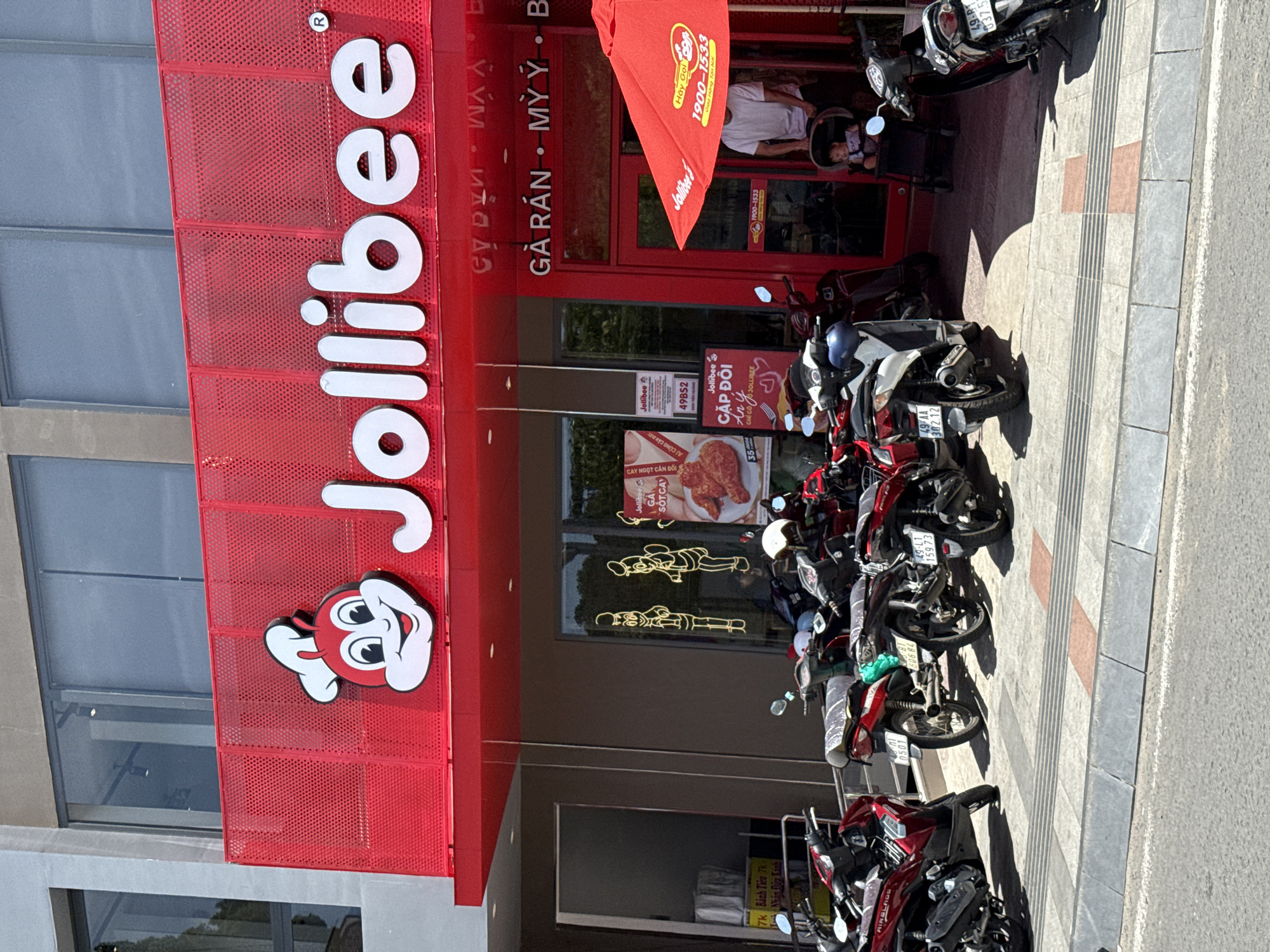 The image shows the exterior of a Jollibee restaurant in Da Lat, Vietnam. The Jollibee signage, in red and white, is prominently displayed above the entrance. Vietnamese text is also on the facade. Motorbikes are parked along the sidewalk in front of the restaurant. The surrounding environment consists of a concrete sidewalk and street, as well as the building structure of the restaurant. A Jollibee advertisement and menu is visible in the restaurant's window, along with figures of Jollibee characters in the window. There is also an advertisement outside the restaurant. A person is seated in the restaurant.